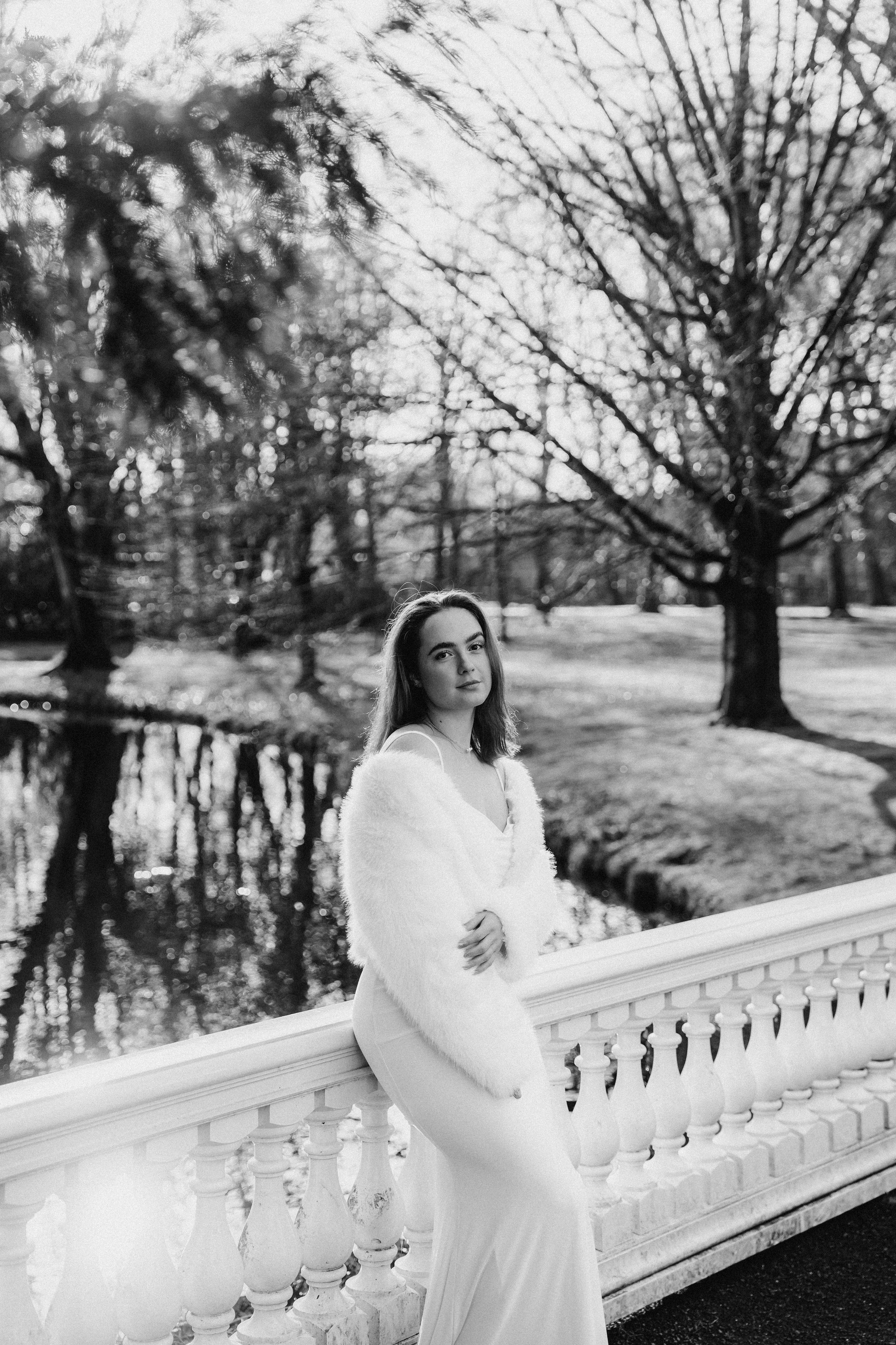 Bruid staat bij een witte balustrade, gekleed in een lange bruidsjurk en een wollen jas, met een park en een meer op de achtergrond, in zwart-wit tijdens een bruidsreportage van Rosa Lynn Signer.