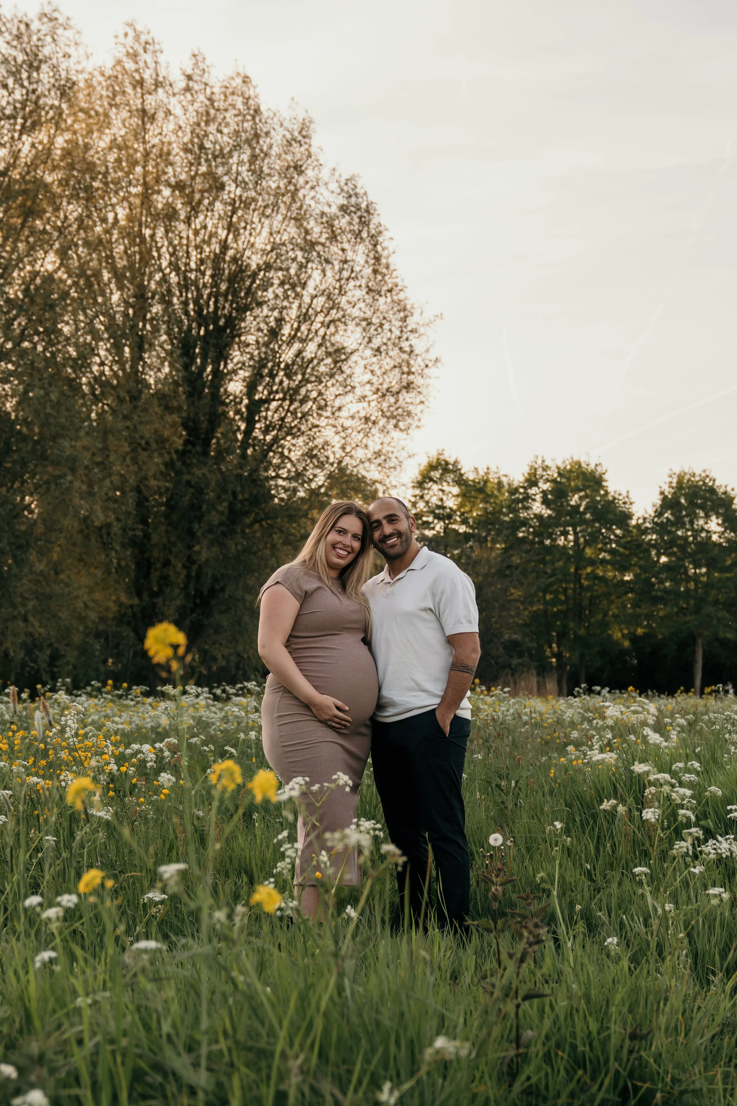 Een gelukkige zwangere vrouw en haar partner staan samen in een bloemrijke veld bij zonsondergang tijdens een zwangerschapsshoot van Rosa Lynn Signer.