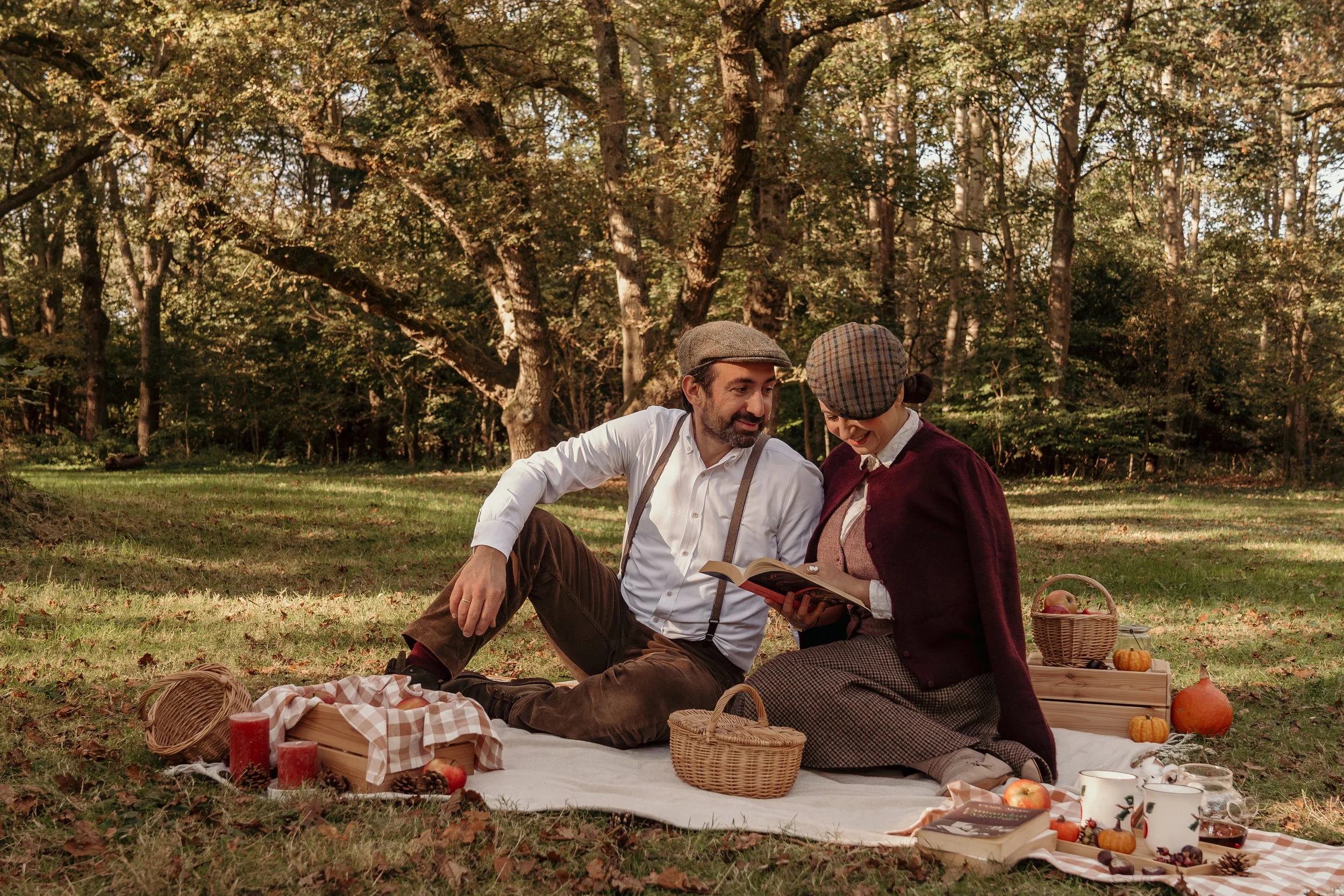 Een man en vrouw zitten samen op een picknickkleed in een bos, genietend van elkaars gezelschap, met herfstdecoraties en eten tijdens een gezinsshoot van Rosa Lynn Signer.