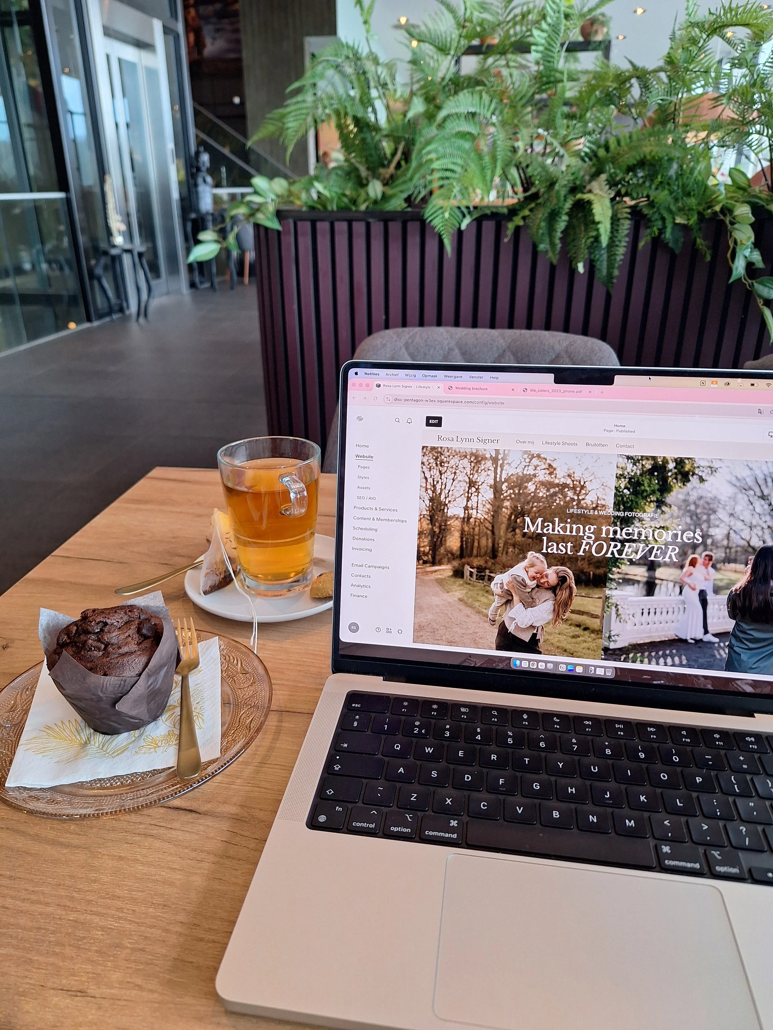 Een laptop op een houten tafel in een cafe, met een kop thee, een muffins, en een vork. Op het scherm is een website zichtbaar met foto's van een bruiloft en de tekst "Making memories last FOREVER". De achtergrond bevat groene planten en grote ramen.