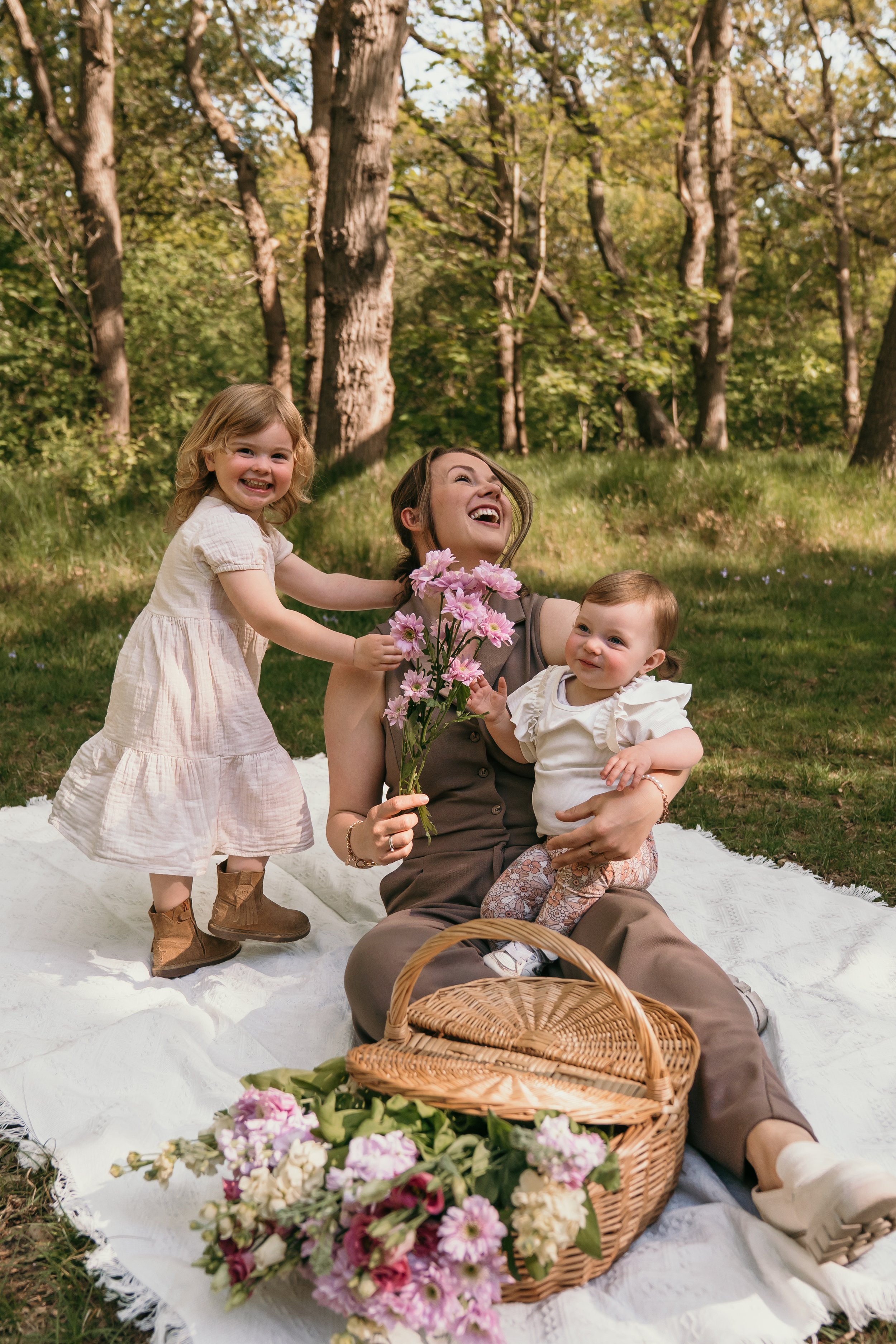 Moeder met haar twee jonge kinderen op een picknick in het bos, bloemen en een mand op een witte deken tijdens mommy & me minishoots van Rosa Lynn Signer. 
