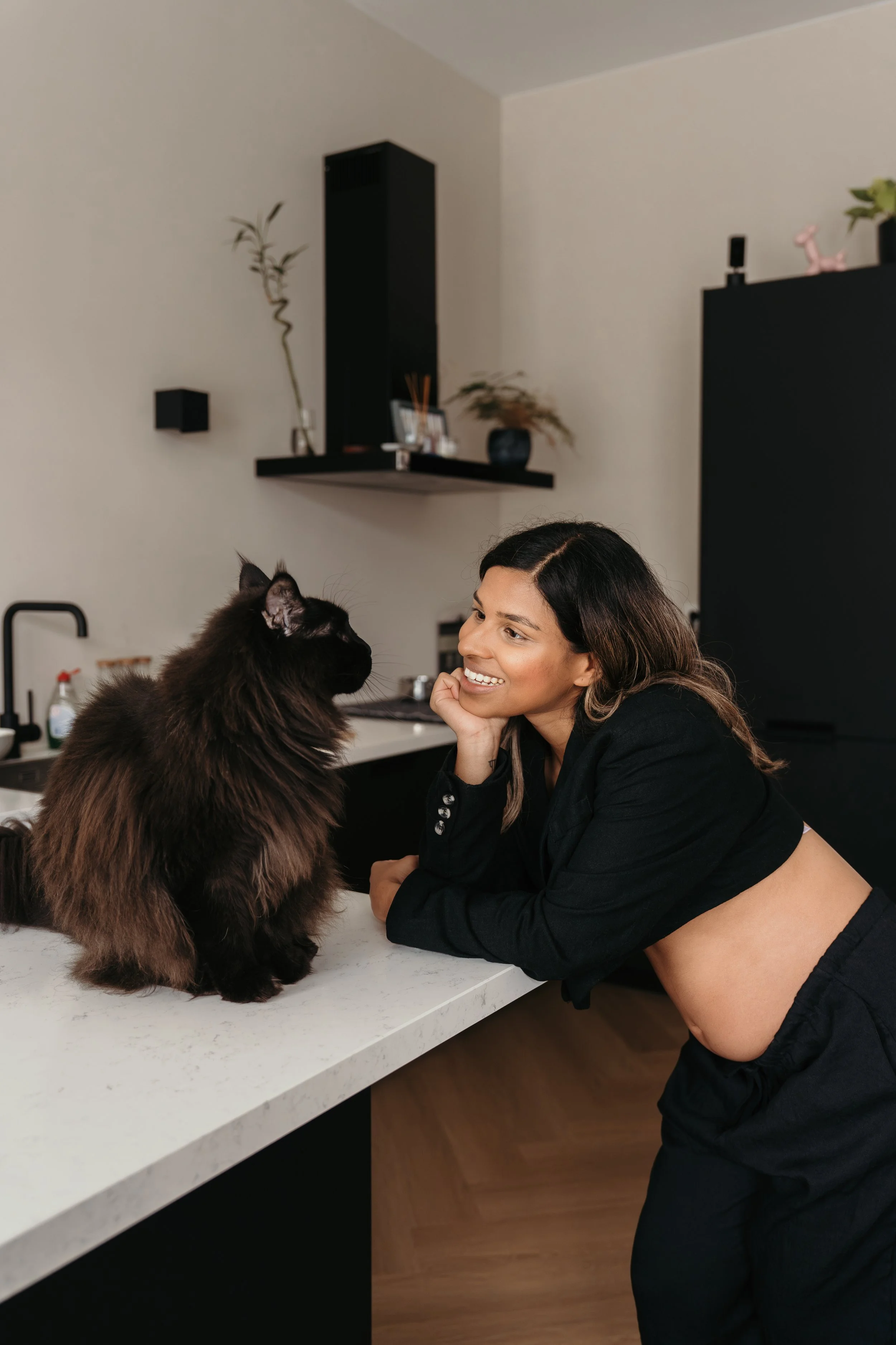 Vrouw met lang haar, glimlachend, leunt op een keuken aanrecht terwijl ze naar een zwarte kat kijkt in een moderne keuken met witte werkblad en zwarte kasten tijdens een zwangerschapsshoot van Rosa Lynn Signer.