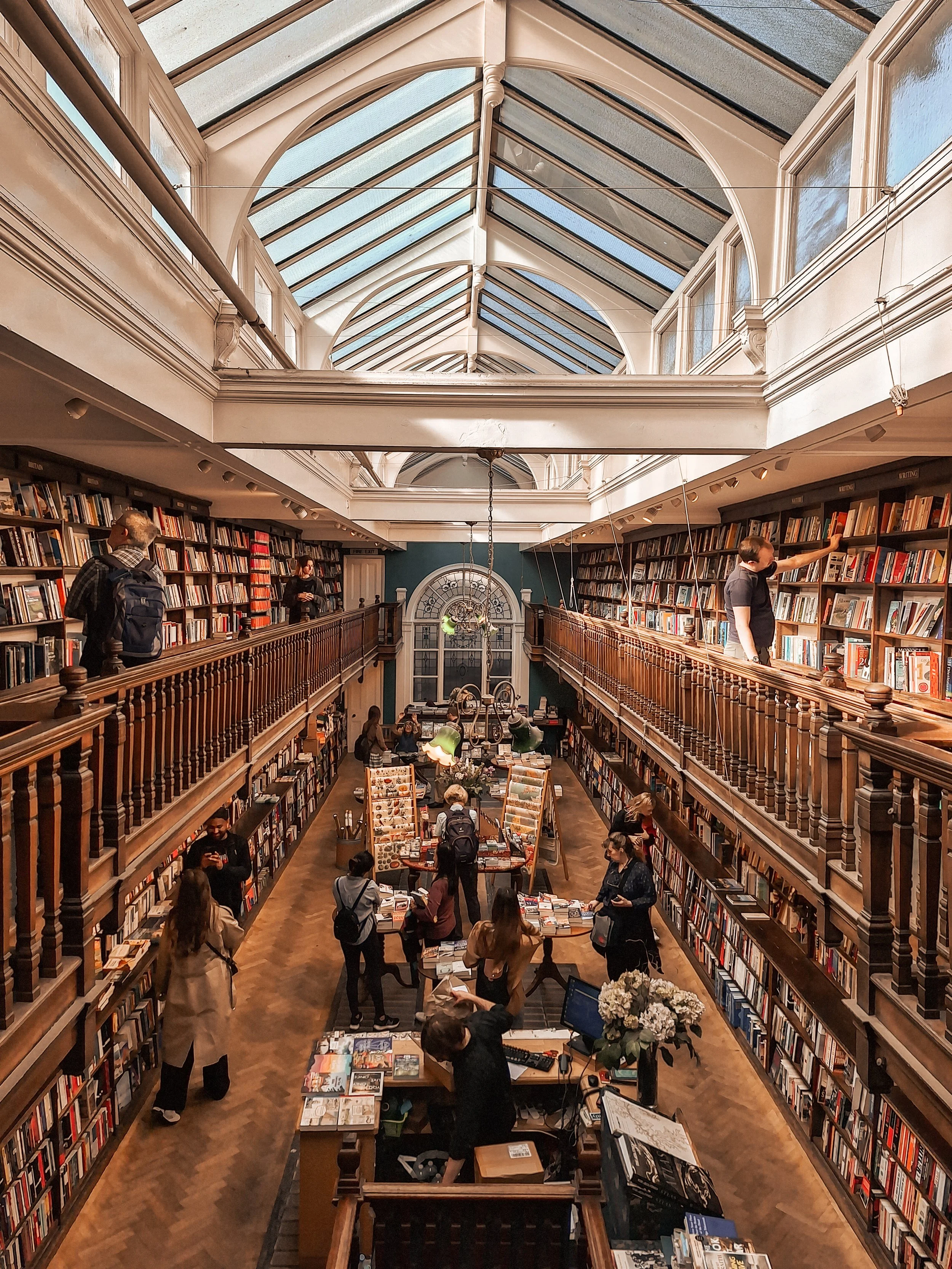 Binnenkant van een oude boekwinkel met hoge houten boekenplanken, een glazen plafond en mensen die boeken bekijken en kopen.