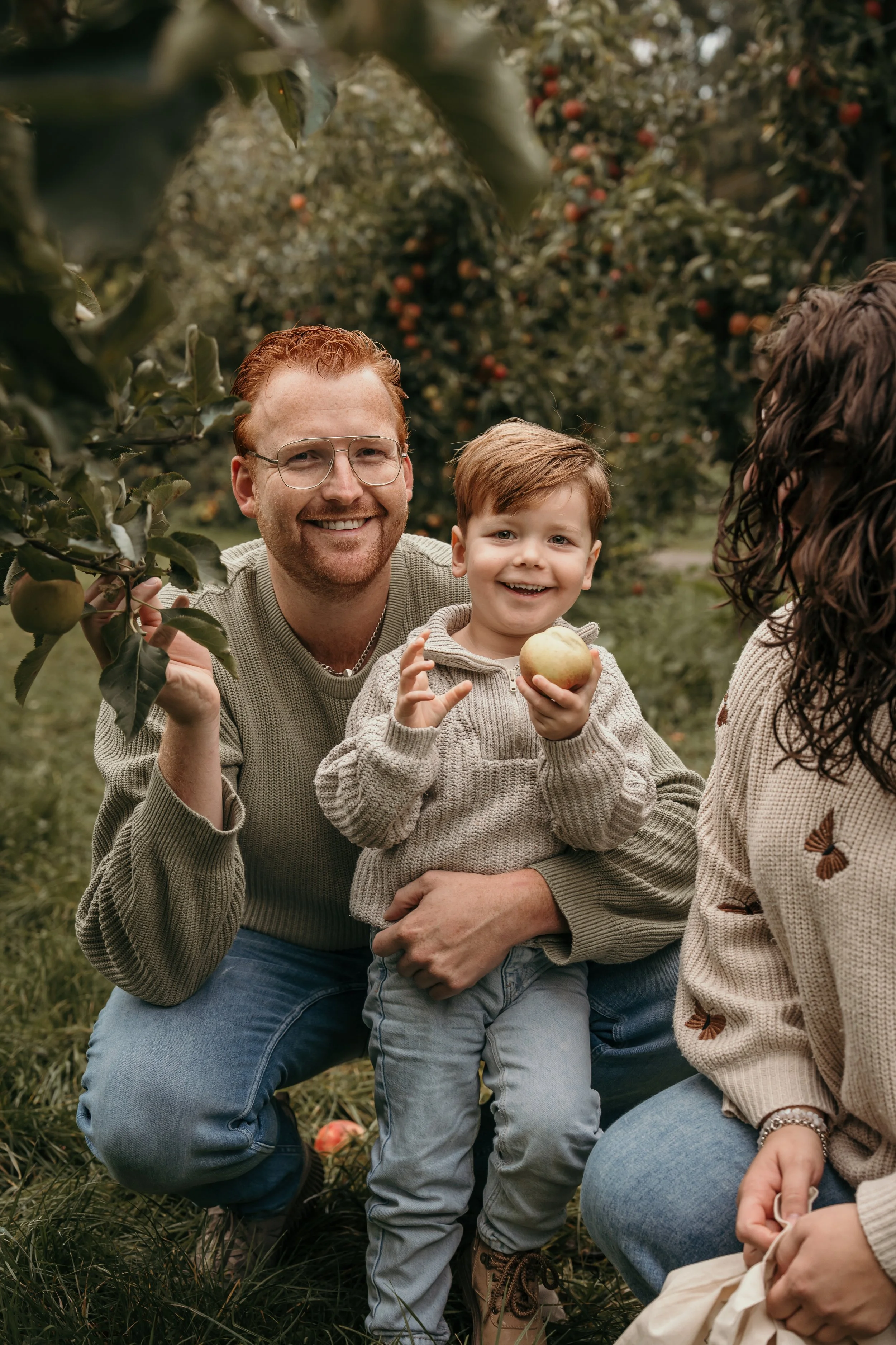Een gelukkige familie in een appelboomgaard, een jongen met een appel, beiden glimlachen tijdens een gezinsshoot van Rosa Lynn Signer.