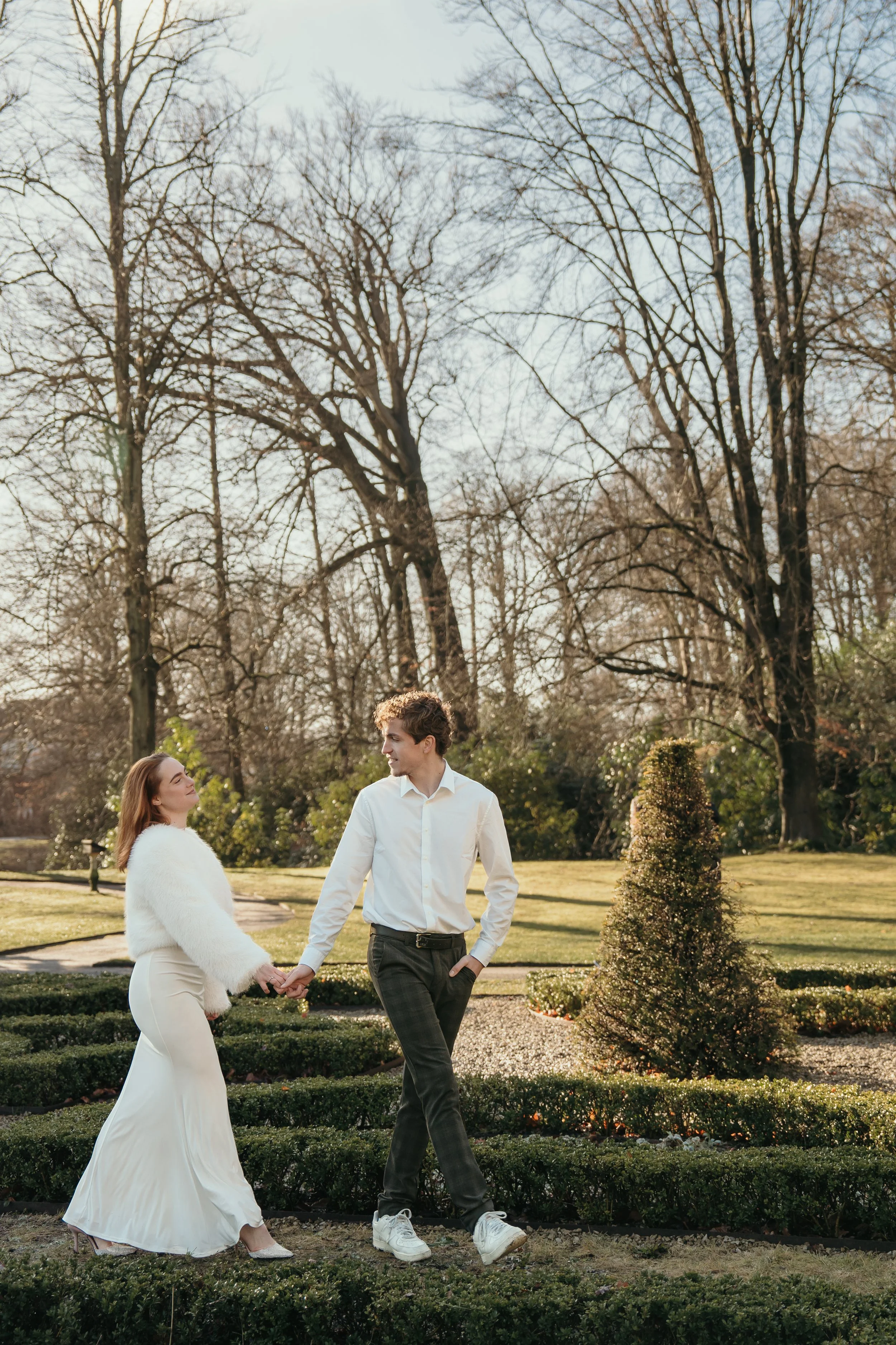 Een jong bruidspaar loopt hand in hand in een park op een zonnige dag, met bomen op de achtergrond tijdens een bruidsreportage van Rosa Lynn Signer.