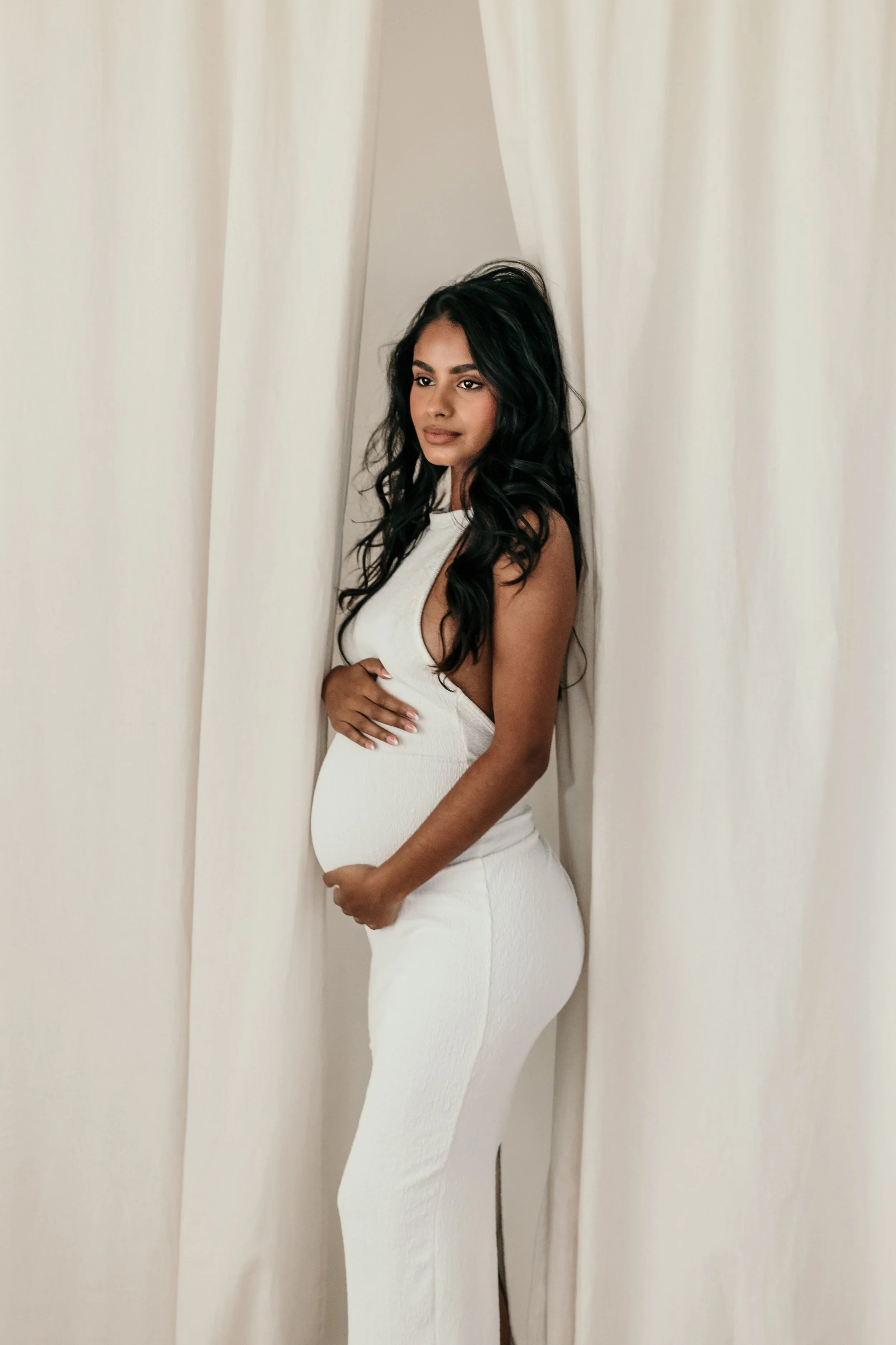 Zwangere moeder met lang donker haar, gekleed in een witte jurk, poseert tussen witte gordijnen tijdens een zwangerschapsshoot van Rosa Lynn Signer in een daglichstudio.