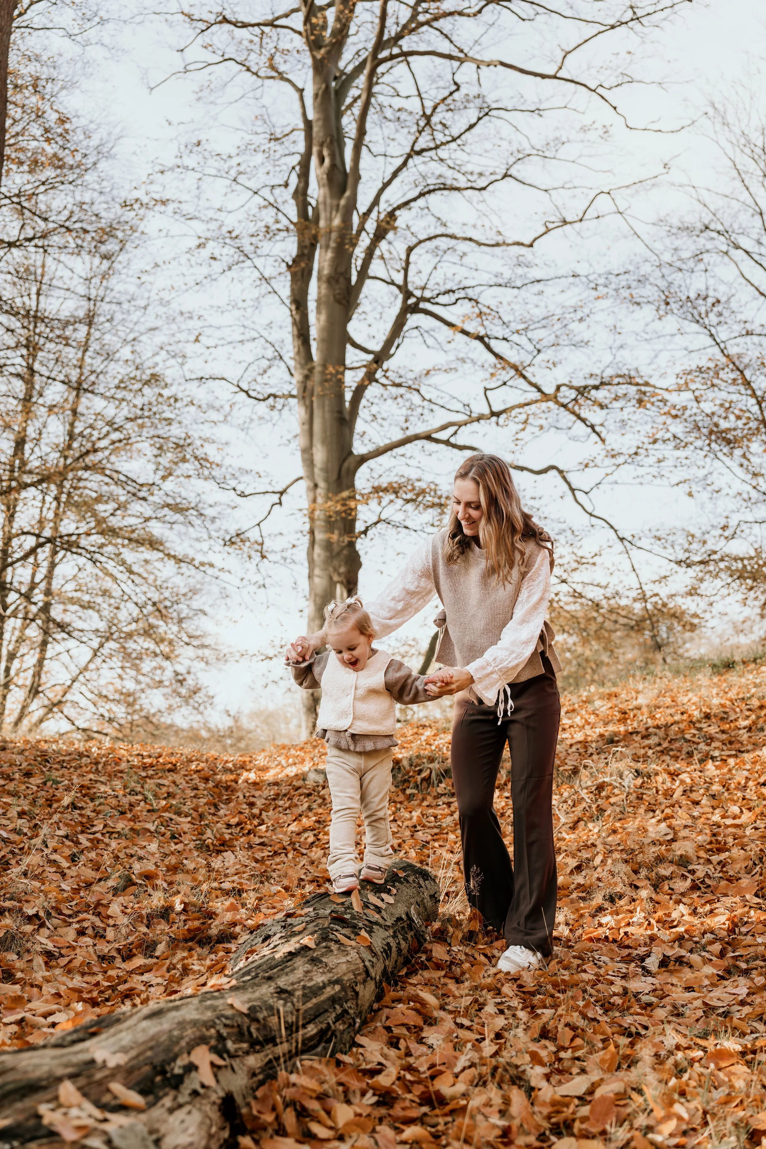 Moeder en dochter lopen over een omgevallen boom in een bos met herfstbladeren en bladverliezende bomen tijdens een moederschap shoot van Rosa Lynn Signer.