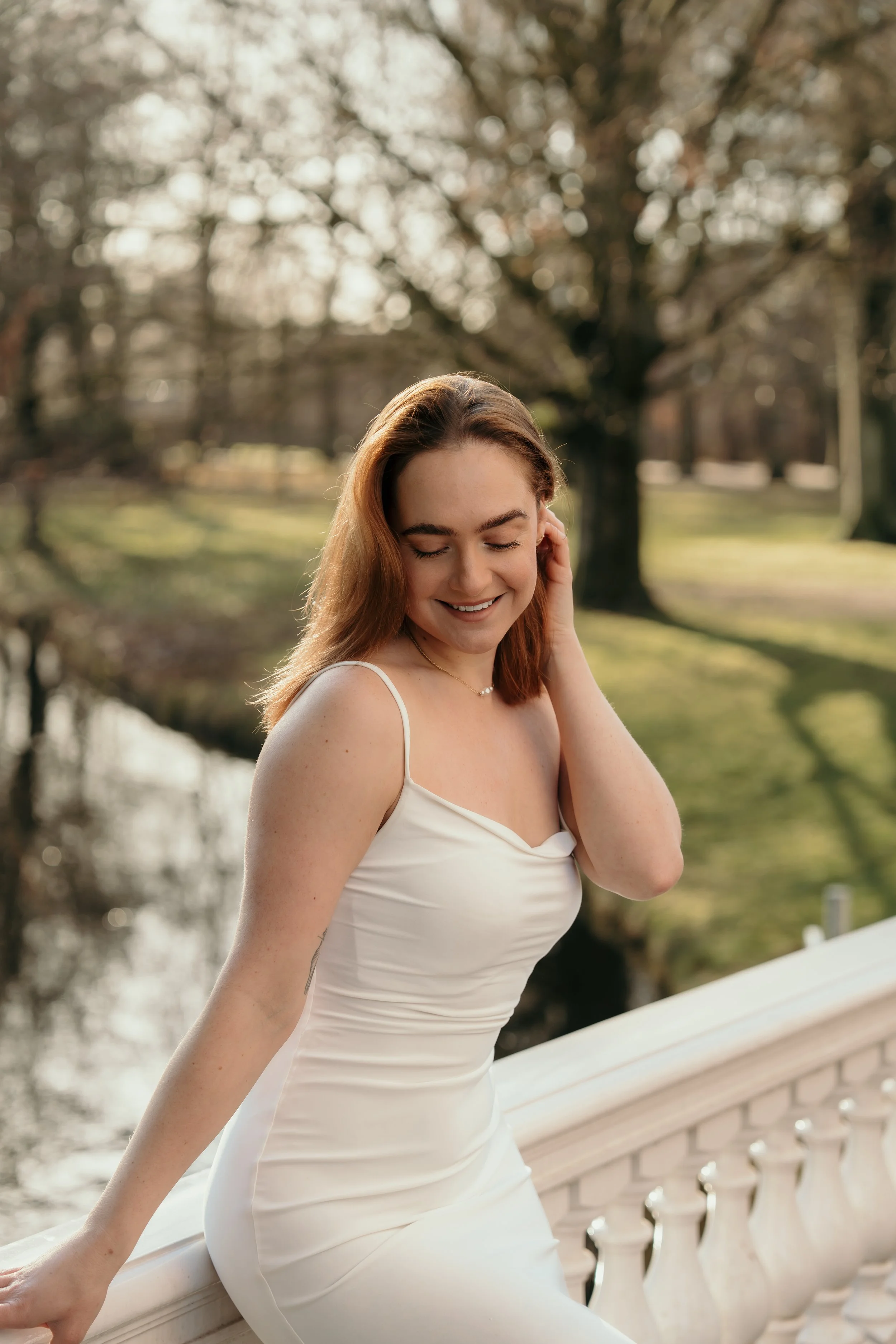 Een lachende bruid in haar trouwjurk staat op een wit balkon met een park en een boom op de achtergrond tijdens een bruidsreportage van Rosa Lynn Signer.
