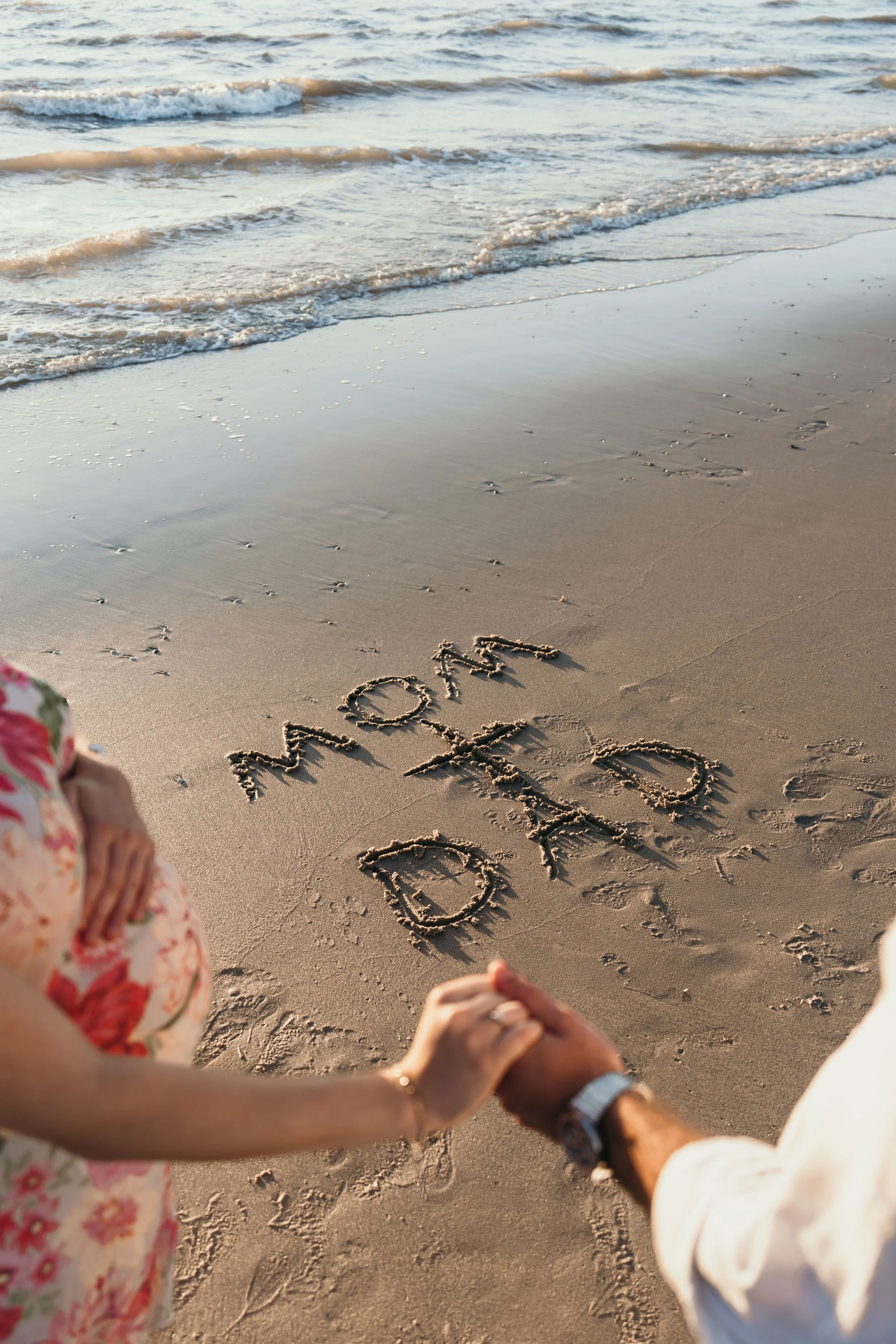 Een koppel die hand in hand op het strand staan met de woorden 'MOM' en 'DAD' in het zand geschreven, met de zee en golven op de achtergrond tijdens een zwangerschapsshoot van Rosa Lynn Signer.