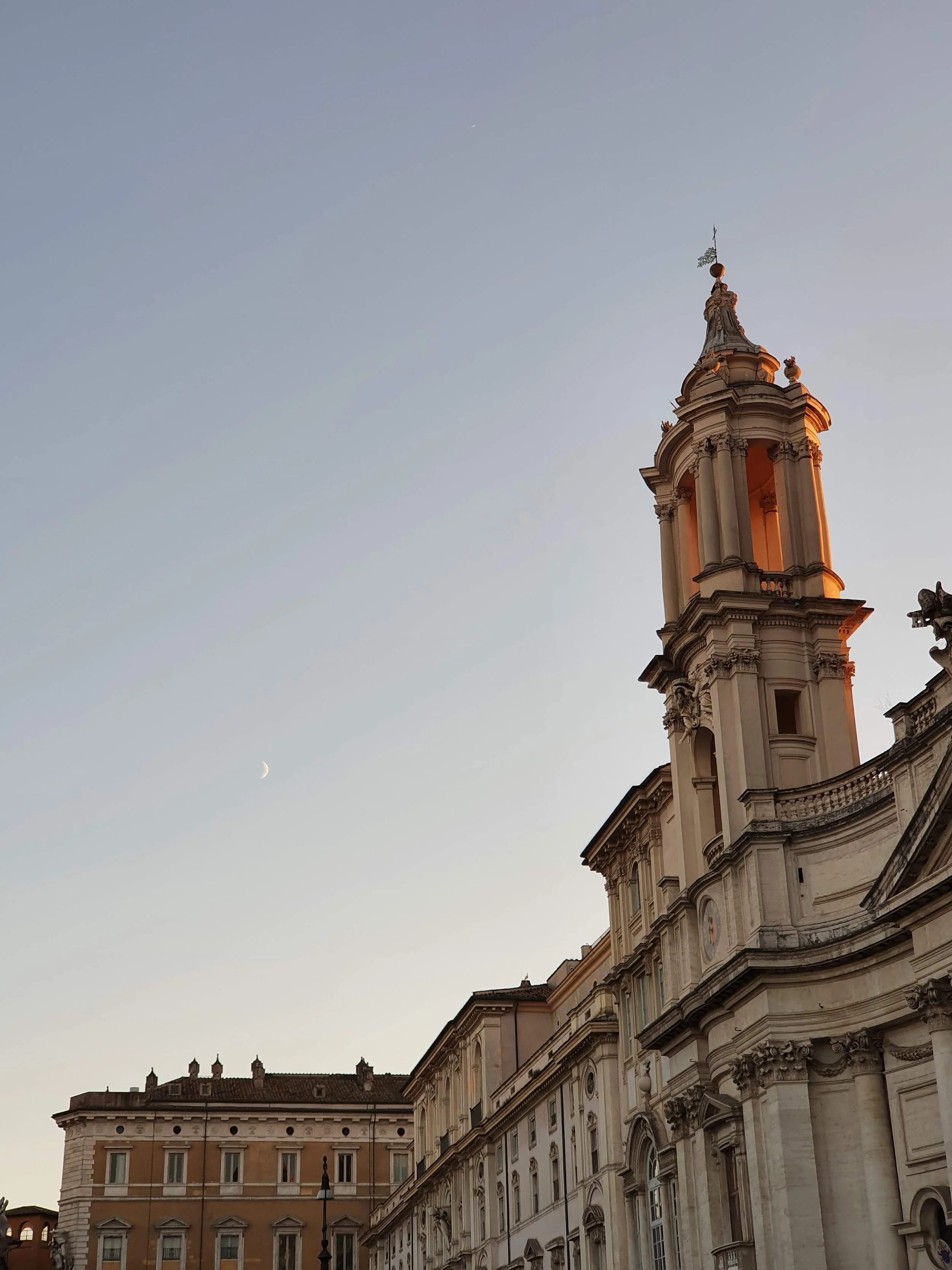 Een barokke kerk of paleis met een toren met een beeld en een weerkaatst zonlicht, onder een heldere hemel met een dunne halve maansikkel.