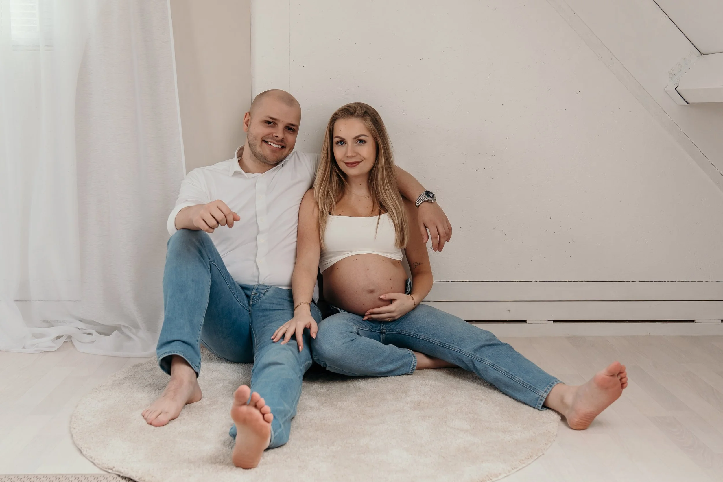 Een zwangere vrouw en een man zitten samen op een tapijt in een lichte kamer, de vrouw heeft haar buik in haar handen en beiden glimlachen tijdens een zwangerschapsshoot van Rosa Lynn Signer in een daglichstudio.