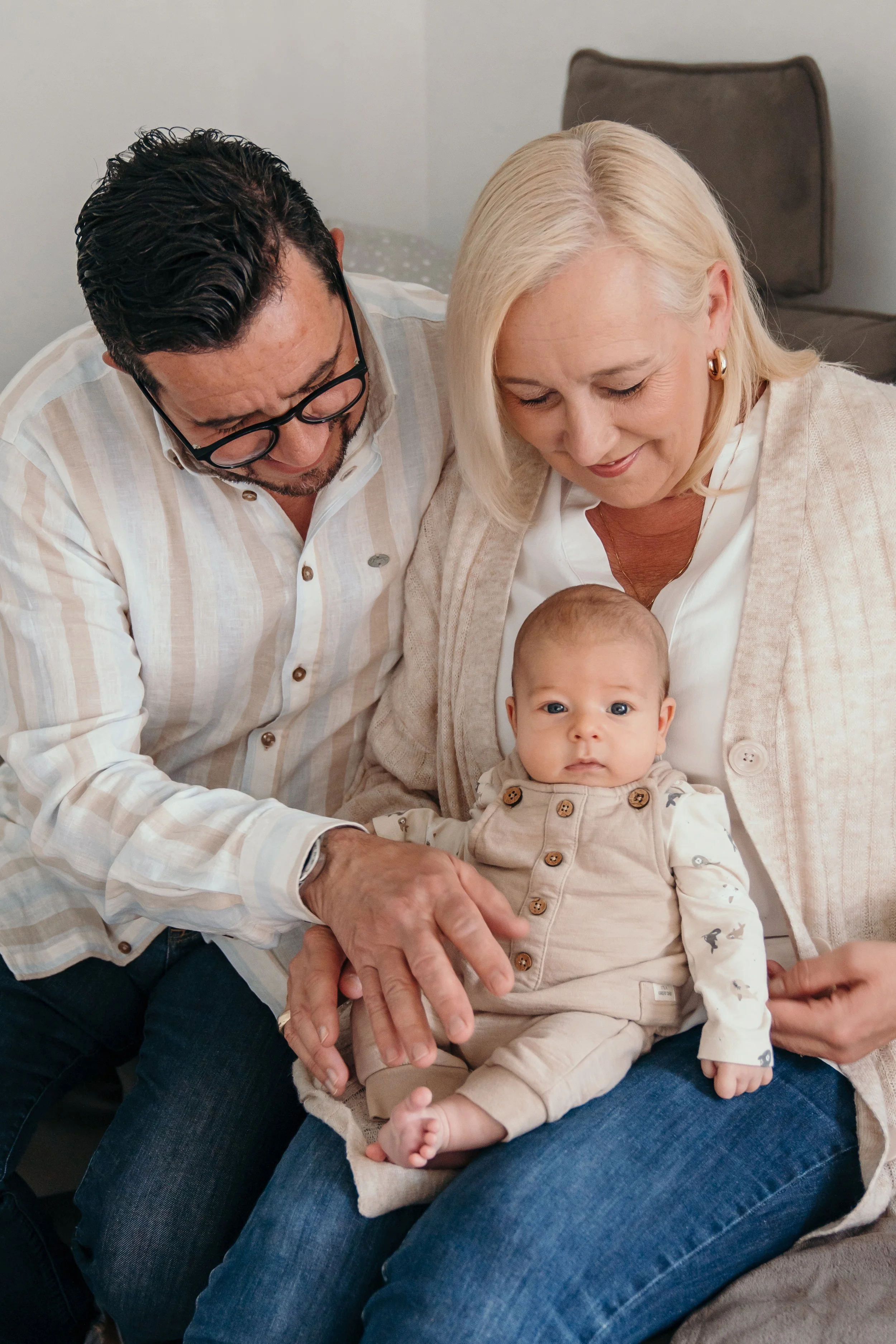 Opa en oma houden new born vast terwijl ze samen zitten op een bank. Opa en oma kijken liefdevol naar de baby, die op haar schoot zit tijdens een new life shoot van Rosa Lynn Signer. 