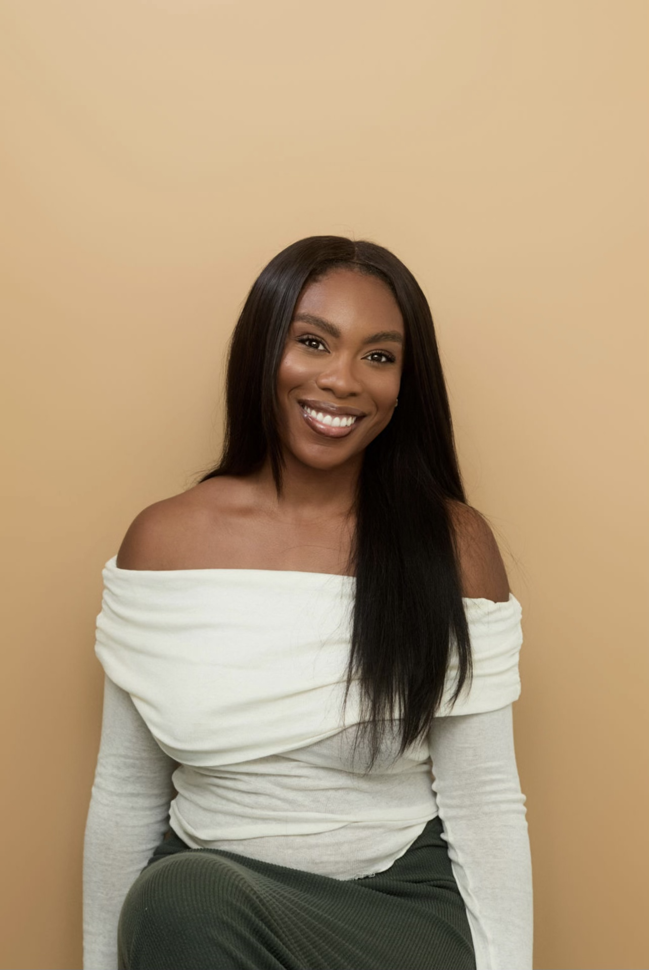 A woman with long black hair, smiling, wearing an off-the-shoulder white top and dark pants, posed against a beige background.