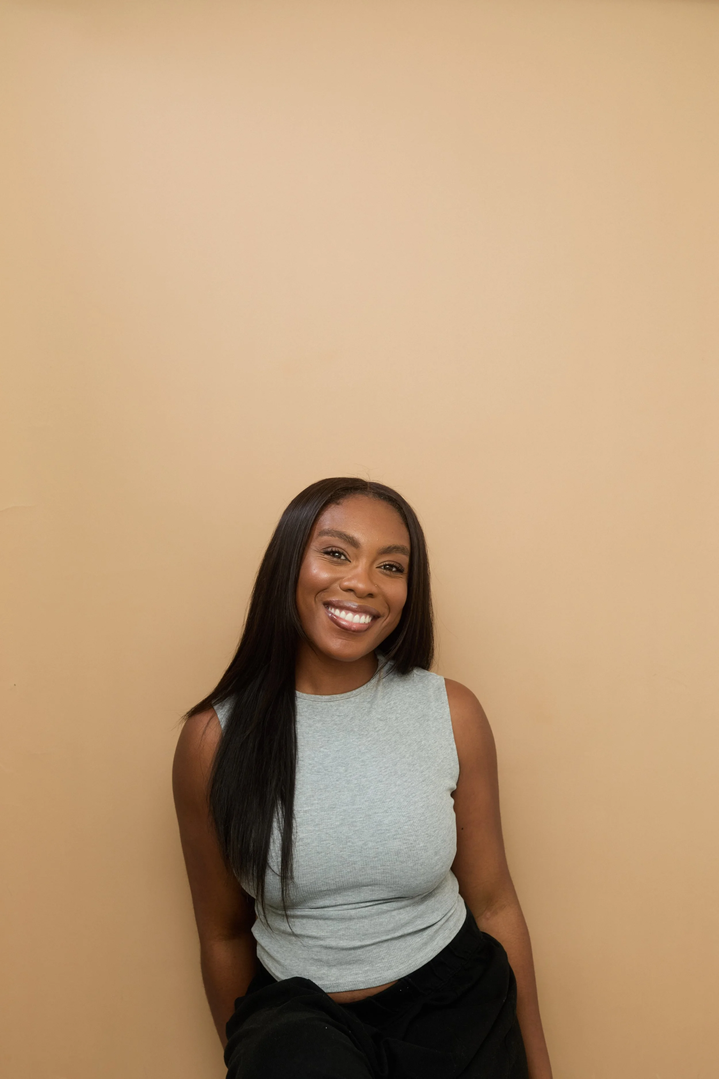 A smiling young woman with long black hair, wearing a sleeveless light gray top and black pants, standing against a beige wall.
