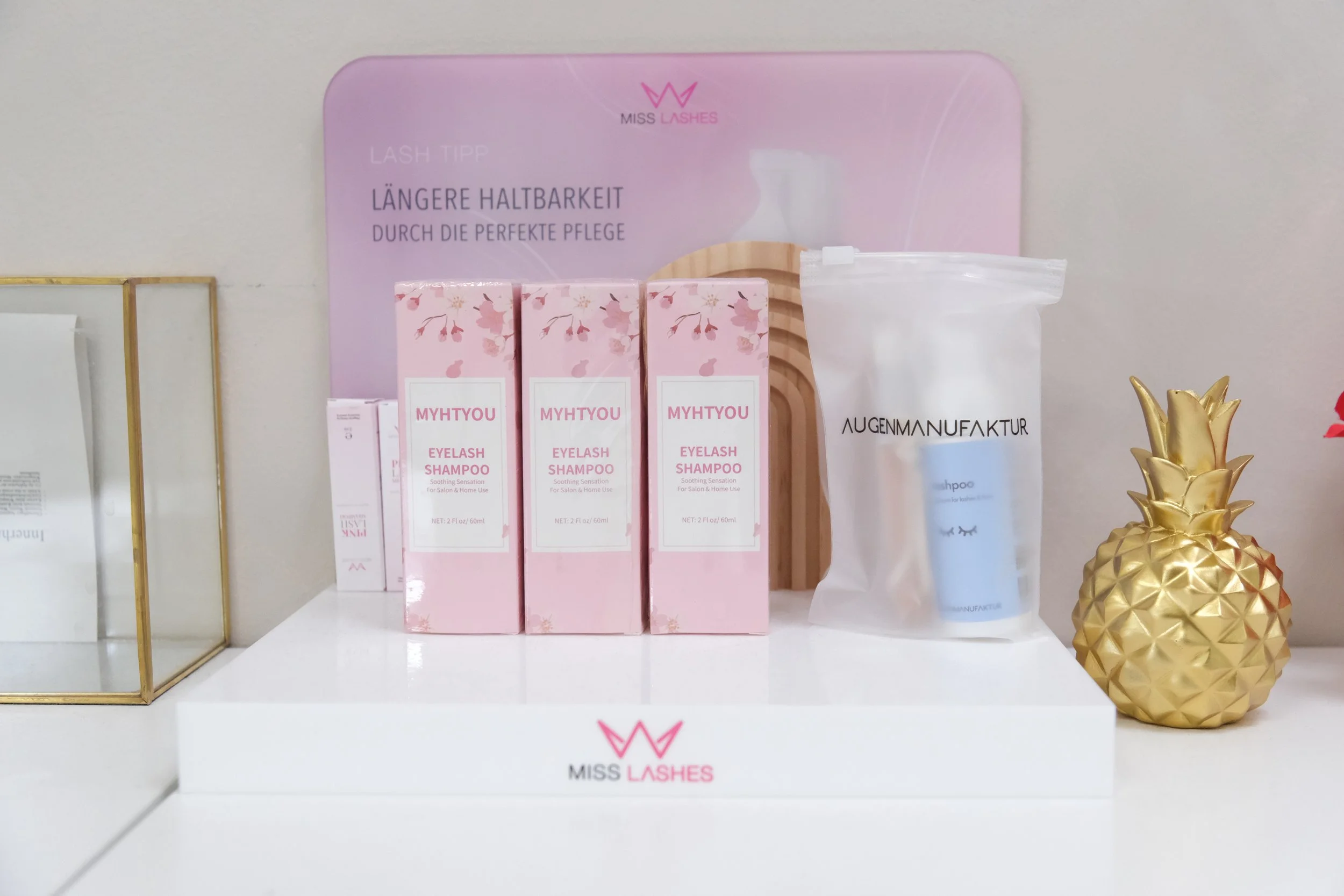Display of pink boxes of MYHTYOU eyelash shampoo, a clear bag labeled AUGENMANUFAKTUR with a blue item inside, a small golden pineapple ornament, and a decorative pink highlight board with German text about eyelash care.