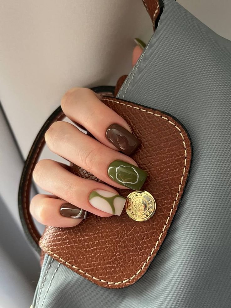 A hand with painted nails, featuring a mix of green and gray nail polish, is holding a small gray bag with a brown leather flap. The flap has a gold button and contrasting white stitching.