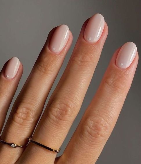 Close-up of a hand with neatly manicured nails painted in a light pink or beige shade, wearing two delicate rings on the fingers.