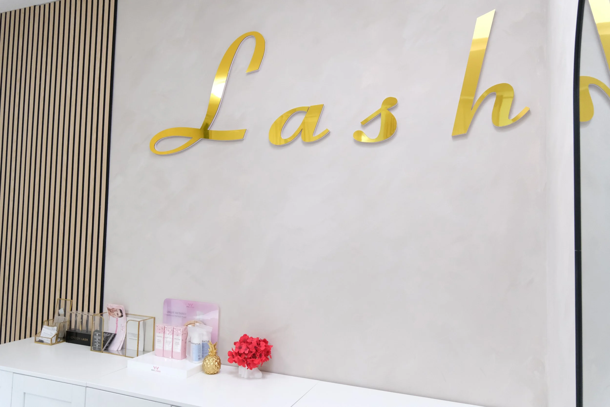 Gold cursive sign spelling 'Lash' on a white wall in a salon or beauty studio, with a white counter below holding beauty products, a small gold pineapple ornament, and a pink flower arrangement.