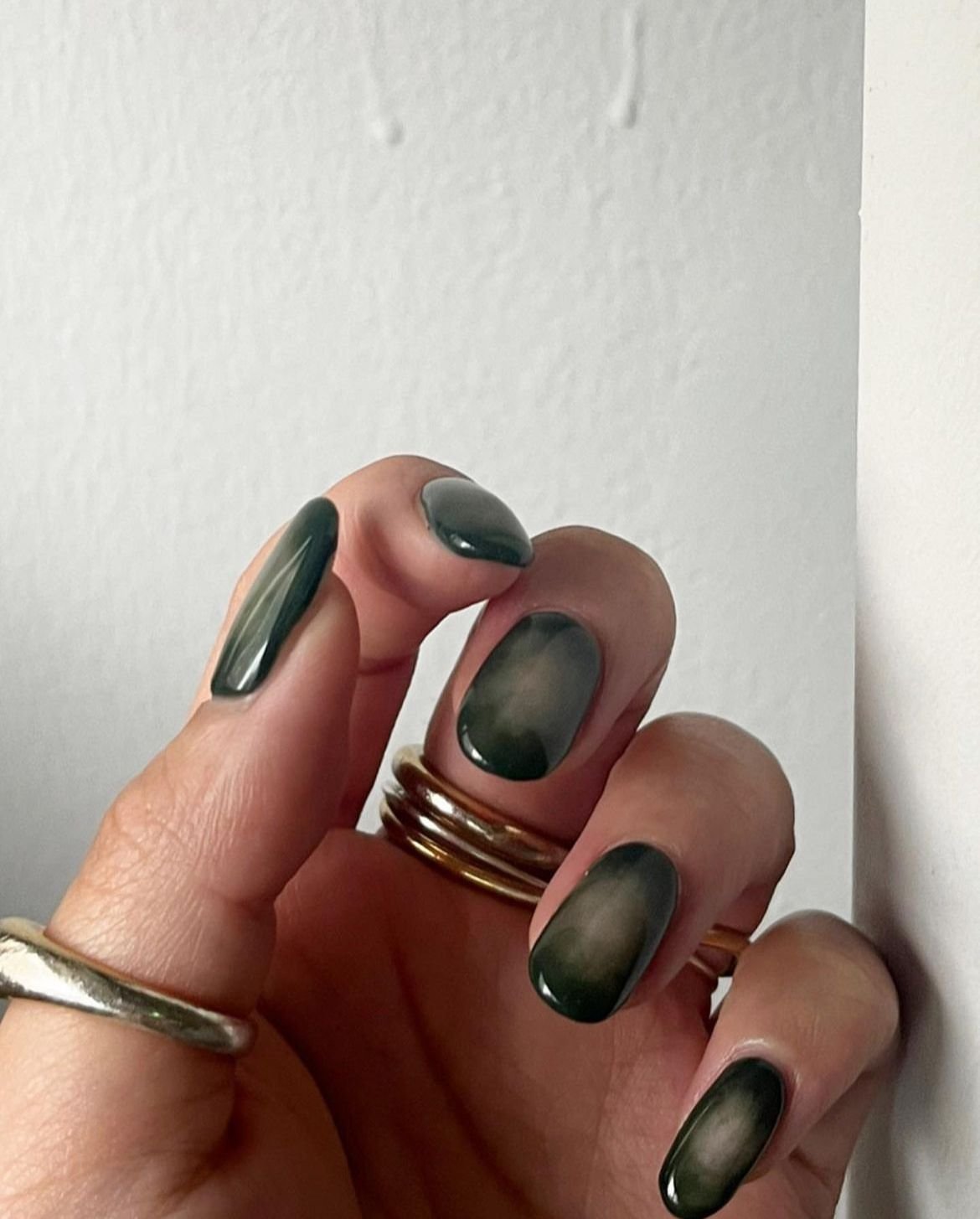 Close-up of a hand with dark, shiny, ombre nail polish, wearing multiple rings against a white wall background.