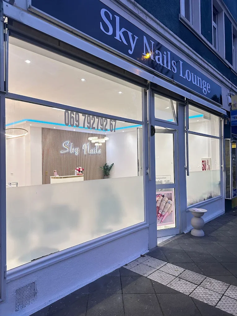 storefront of Sky Nails Lounge with large windows displaying the interior, a white reception desk, a neon sign reading Sky Nails, potted plant, and a promotional sign indicating 30% off; the exterior features a tactile paving strip on the sidewalk.