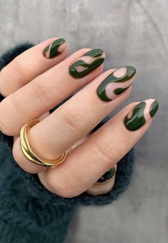 Close-up of a hand with green and beige patterned gel nails and wearing a gold ring, against a gray background.