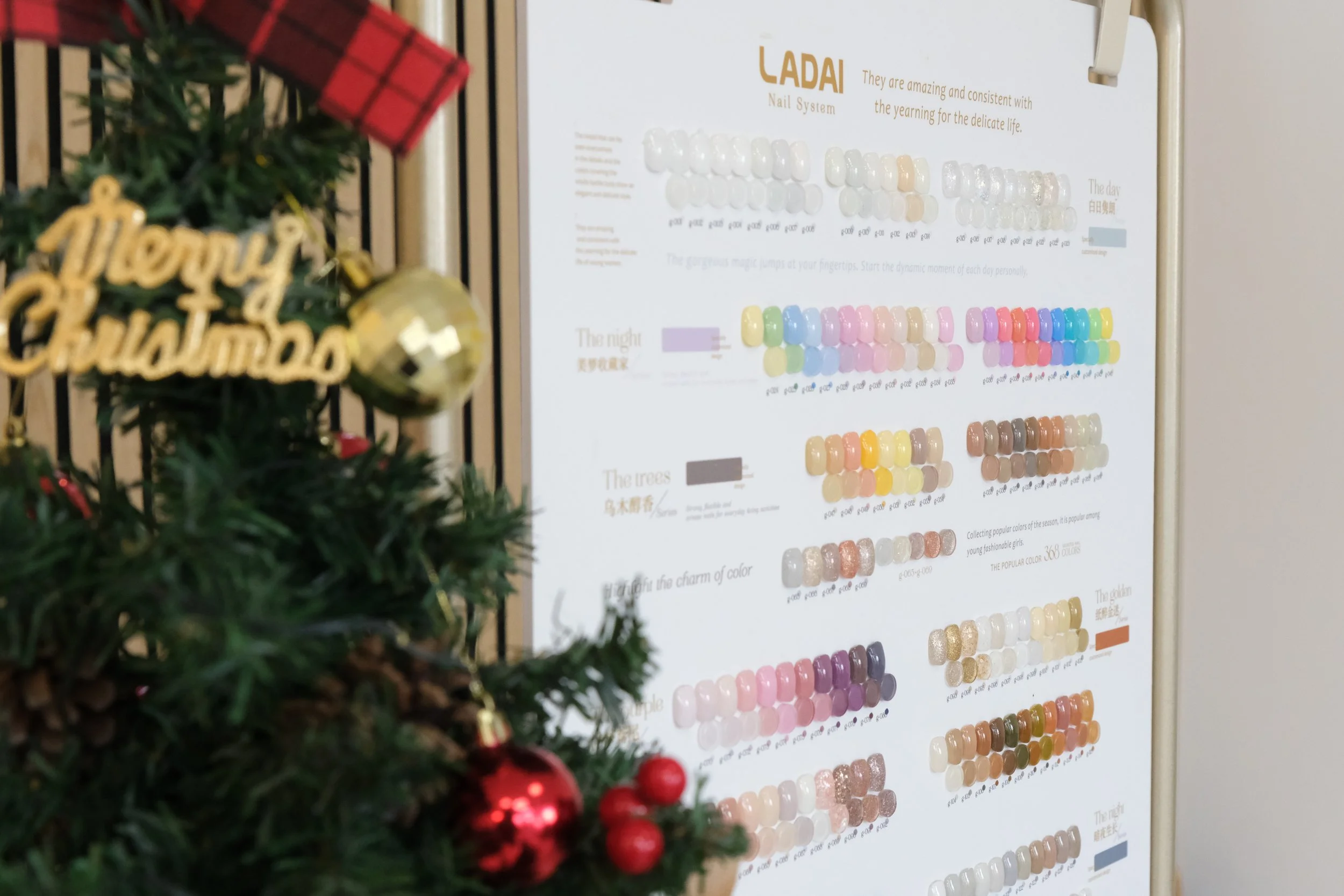 Part of a decorated Christmas tree with a gold ornament and red baubles, and a sign that says "Merry Christmas" in gold. In the background, a nail color chart display with various color samples and descriptions.