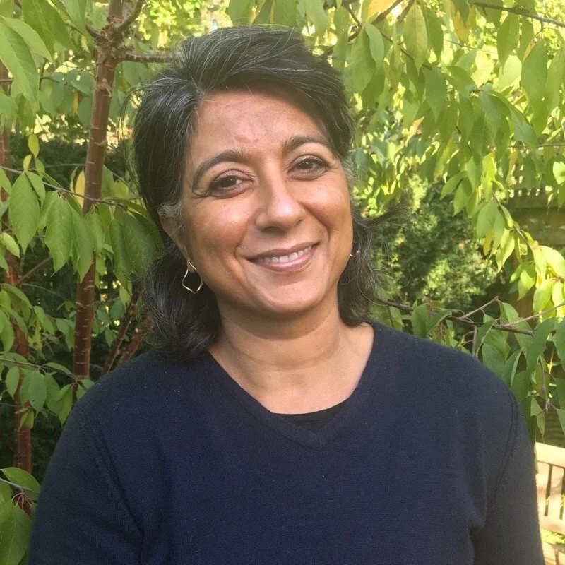 Della K Gould: A counsellor and therapist with dark hair and earrings smiling outdoors surrounded by green leaves.