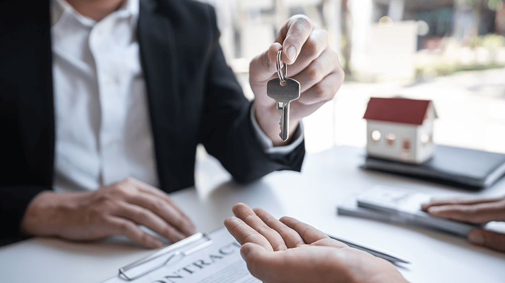Person in suit handing over key to another person during a real estate contract signing, with small house model in background.