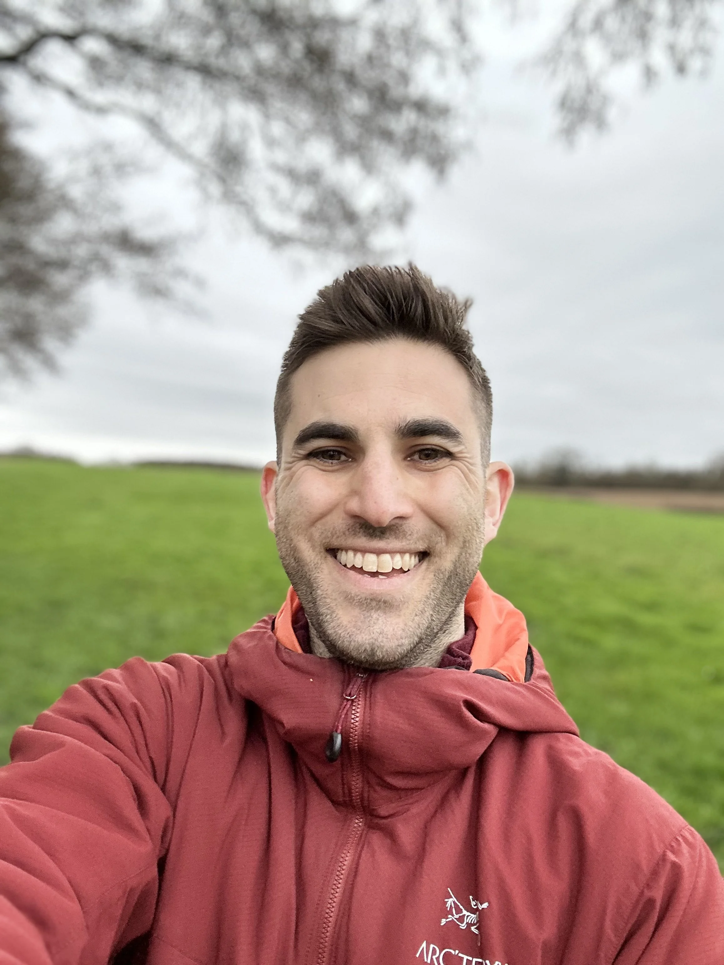 Dr Matt Slavin, Clinical Psychologist smiling taking a selfie outdoors on a cloudy day, with green grass and trees in the background, wearing a red Arc'teryx jacket.