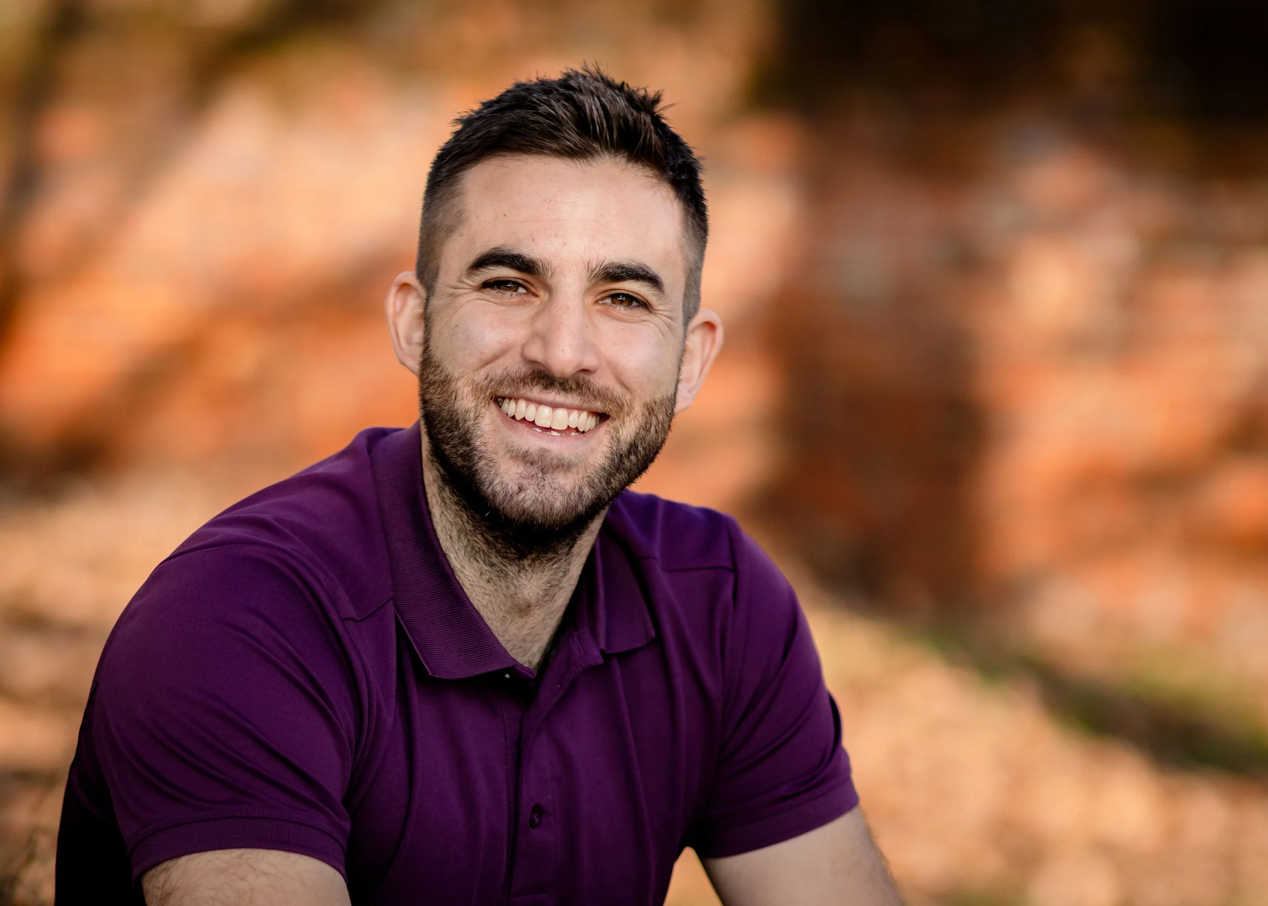 Dr Matt Slavin, Clinical Psychologist with short dark hair and a beard, wearing a purple polo shirt, outdoors during fall with blurred orange and brown leaves in the background.