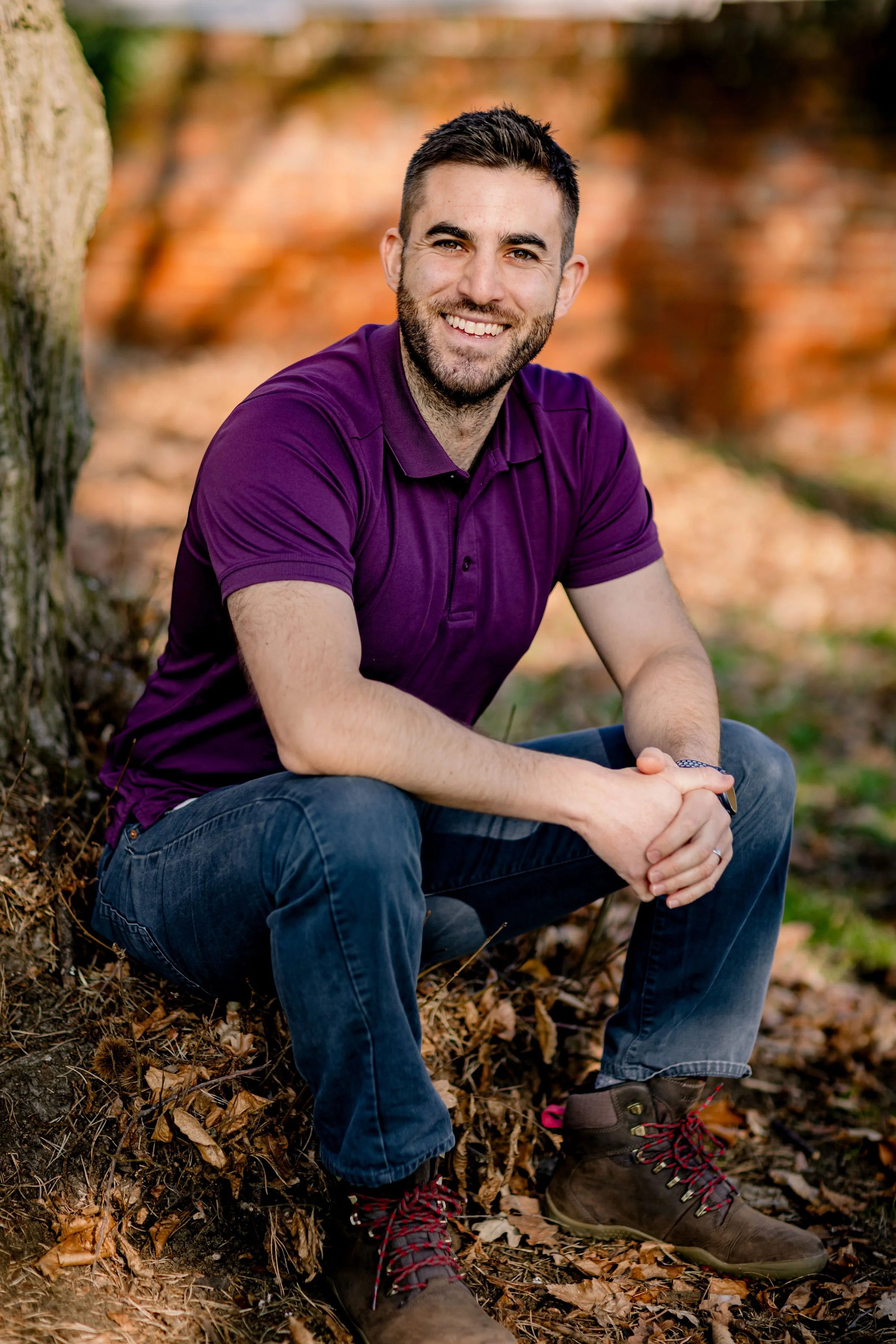 Dr Matt Slavin, Clinical Psychologist  itting outdoors on autumn leaves, smiling, wearing a purple polo shirt, jeans, and hiking boots, with a blurred fall-colored background.