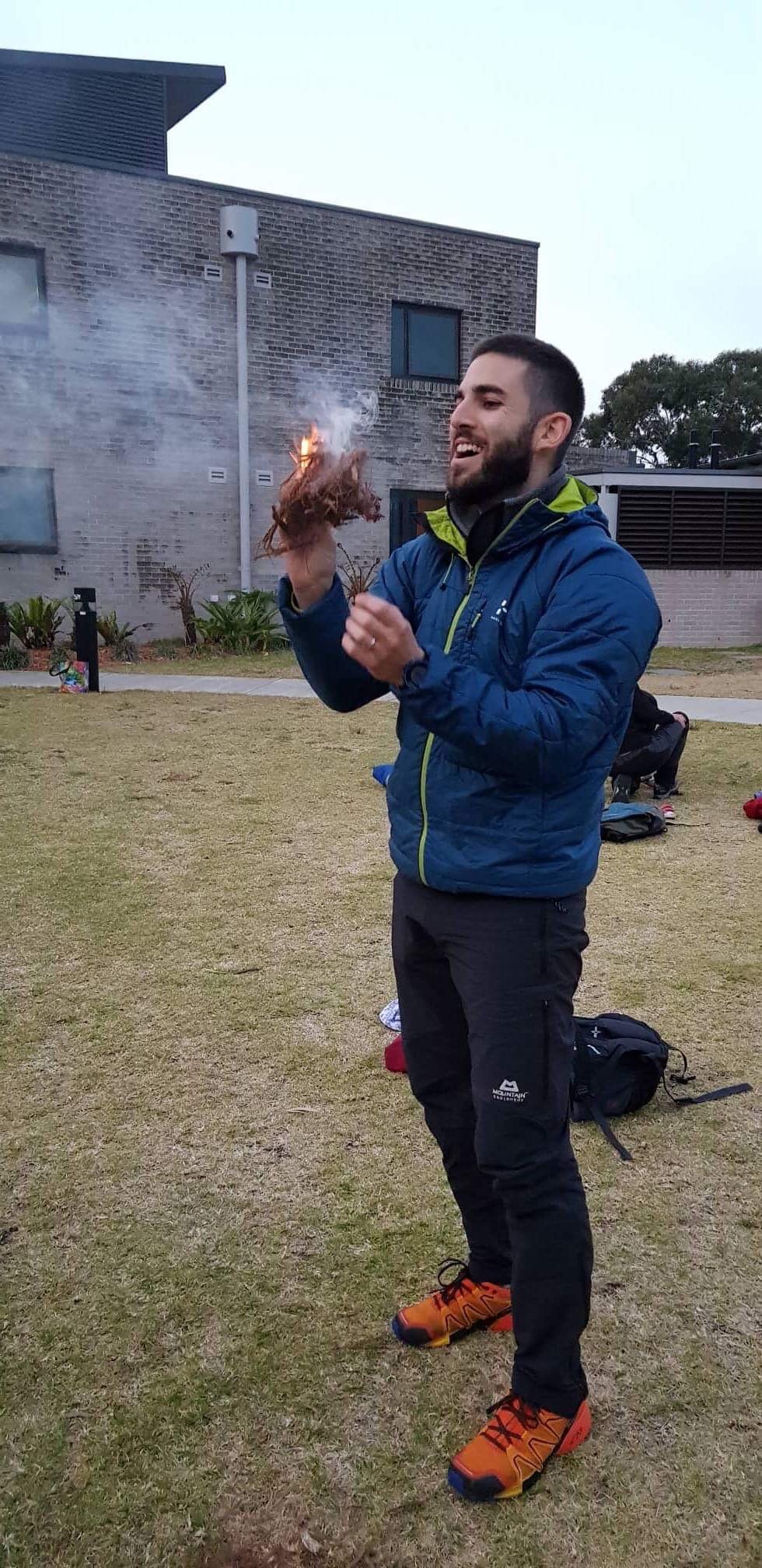 Dr Matt Slavin, Clinical Psychologist outdoors in a grassy area holding a small burning object, smiling. He is wearing a blue jacket, black pants, and orange shoes, with a backpack on the ground nearby.