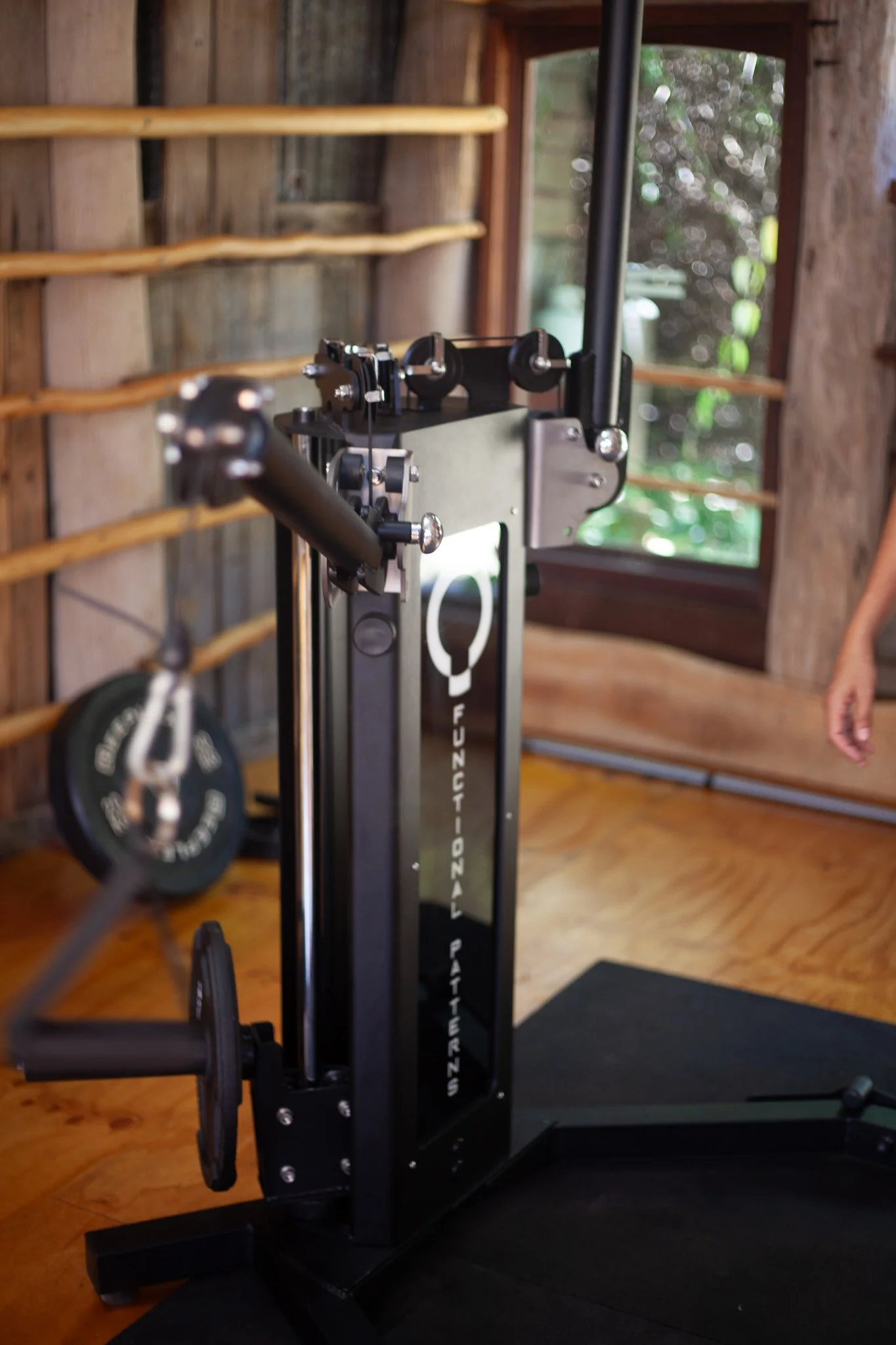 A piece of fitness equipment inside a rustic wooden room with a window and weight plates in the background.