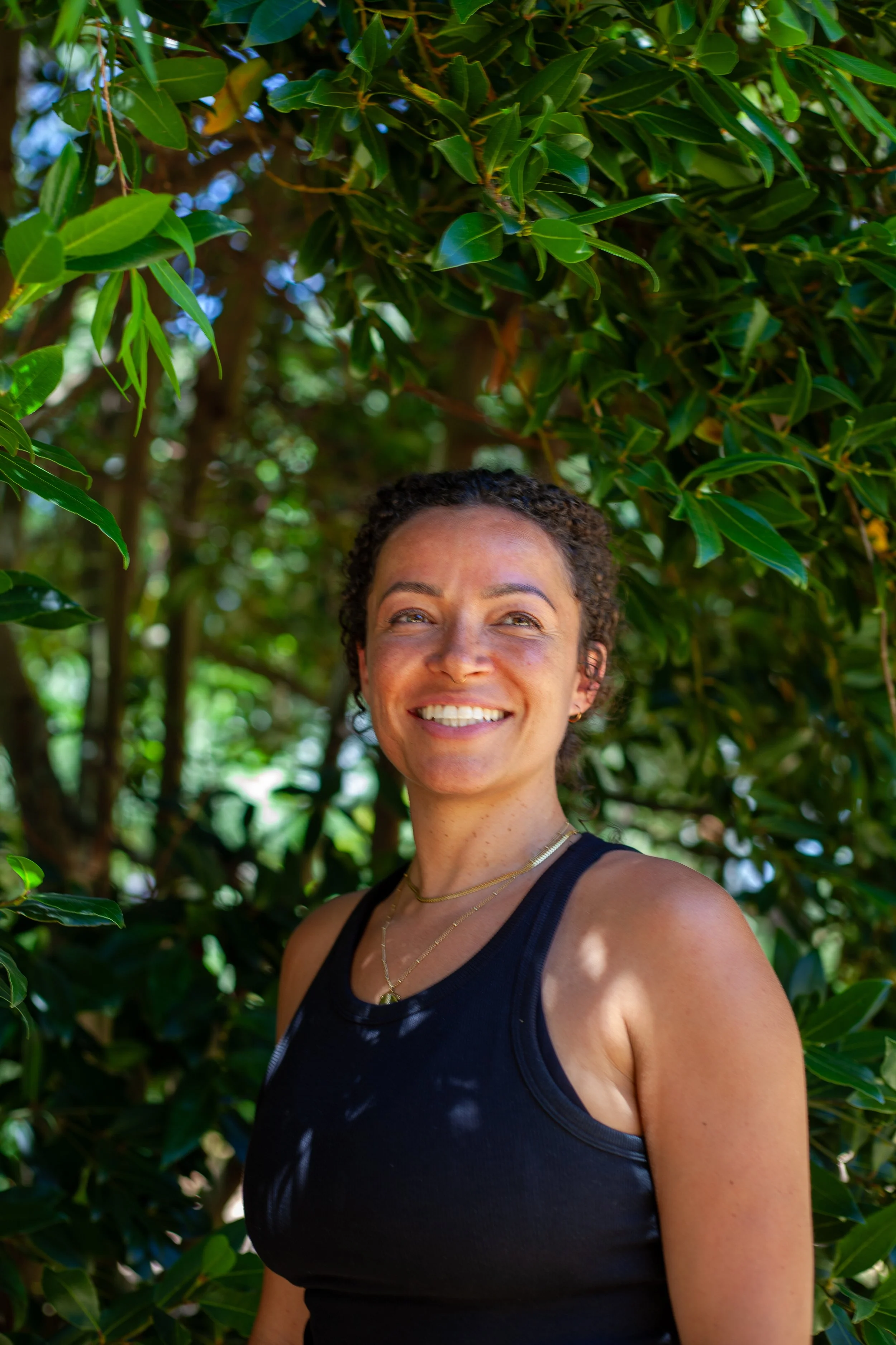 Smiling woman with curly hair wearing a black tank top and gold jewelry, standing outdoors among green leafy bushes.