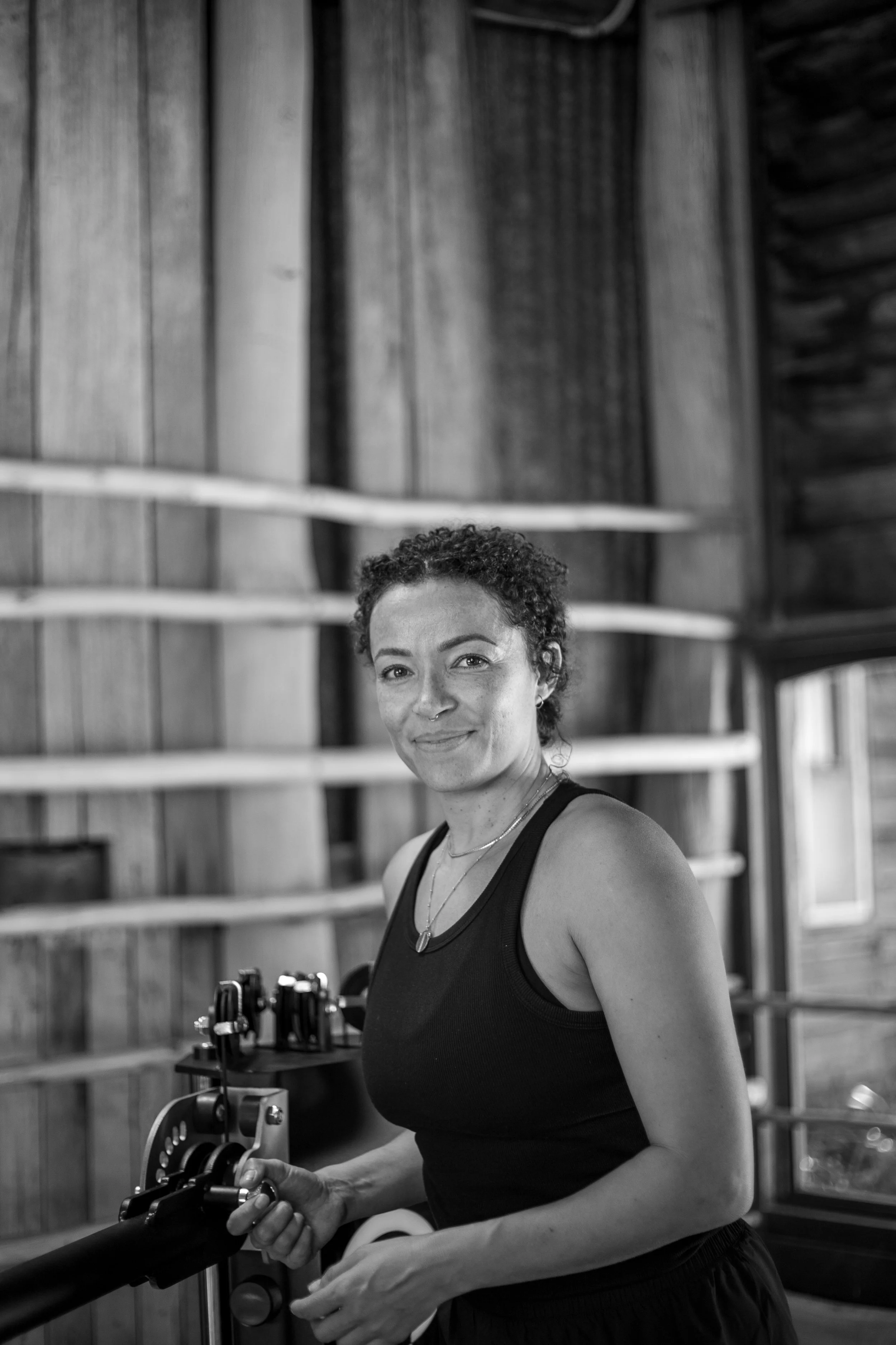 A woman with short curly hair and a nose piercing, wearing a sleeveless black top, stands indoors in front of wooden shelves with exercise equipment, smiling at the camera.