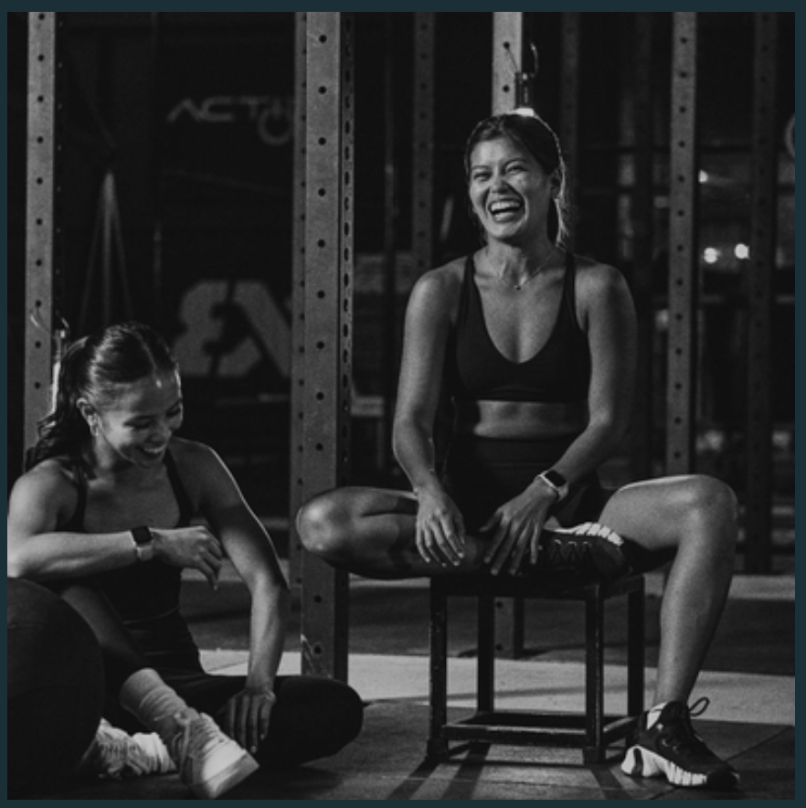 Two women in workout clothing smiling and laughing in a gym. One woman is sitting on a stool, and the other is sitting on the floor, tying her shoelace.