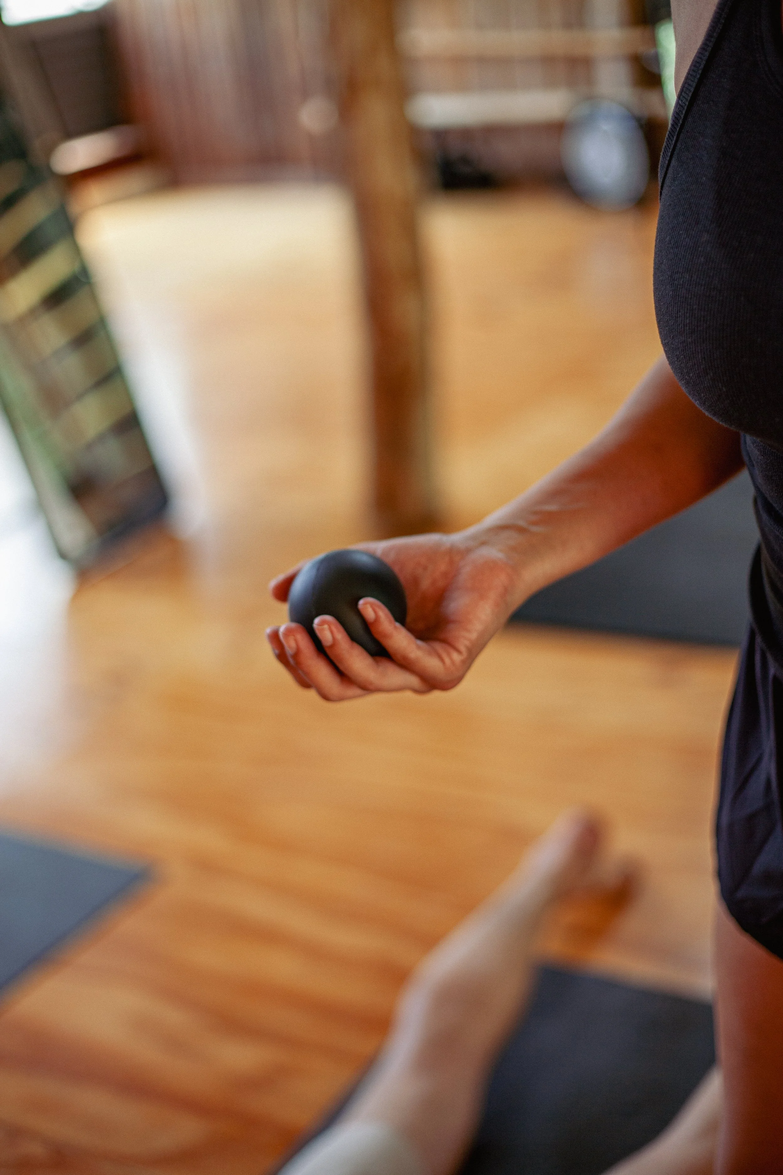 Person holding a black massage roller while another person lies on a yoga mat on wooden flooring.