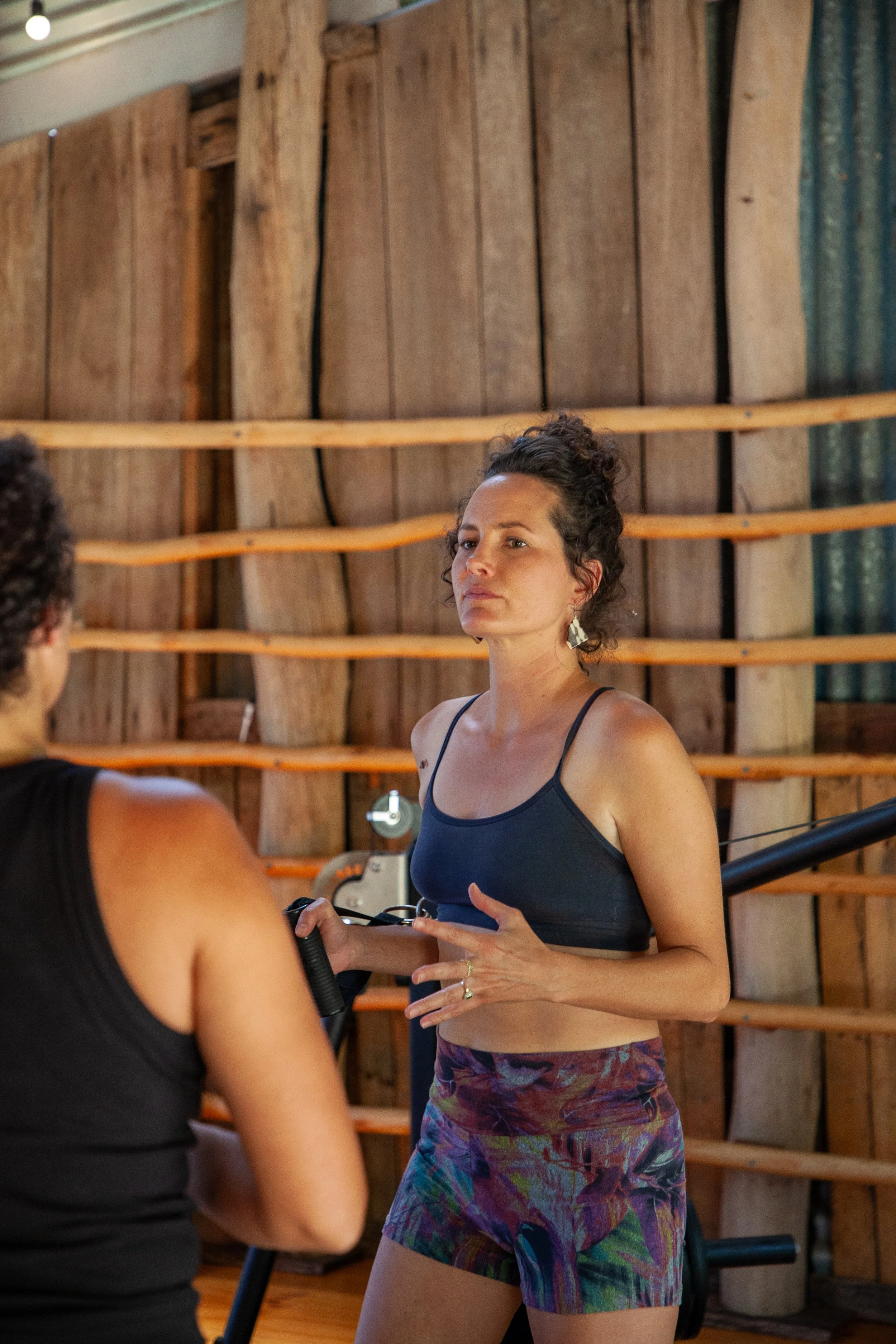 Two women having a conversation in a rustic wooden gym. One woman is wearing a black sports bra and colorful shorts, and the other, whose face is not visible, is wearing a black tank top.