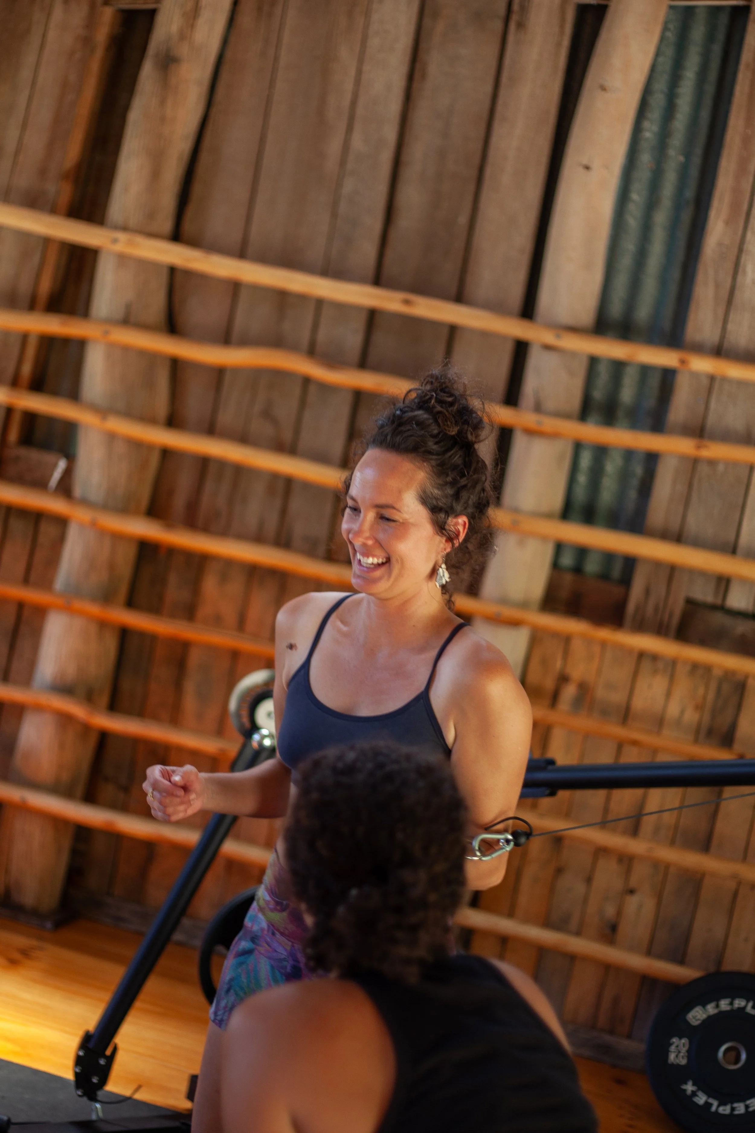 Two women participating in a fitness class inside a wooden gym, one standing and smiling, the other seated with her back to the camera.