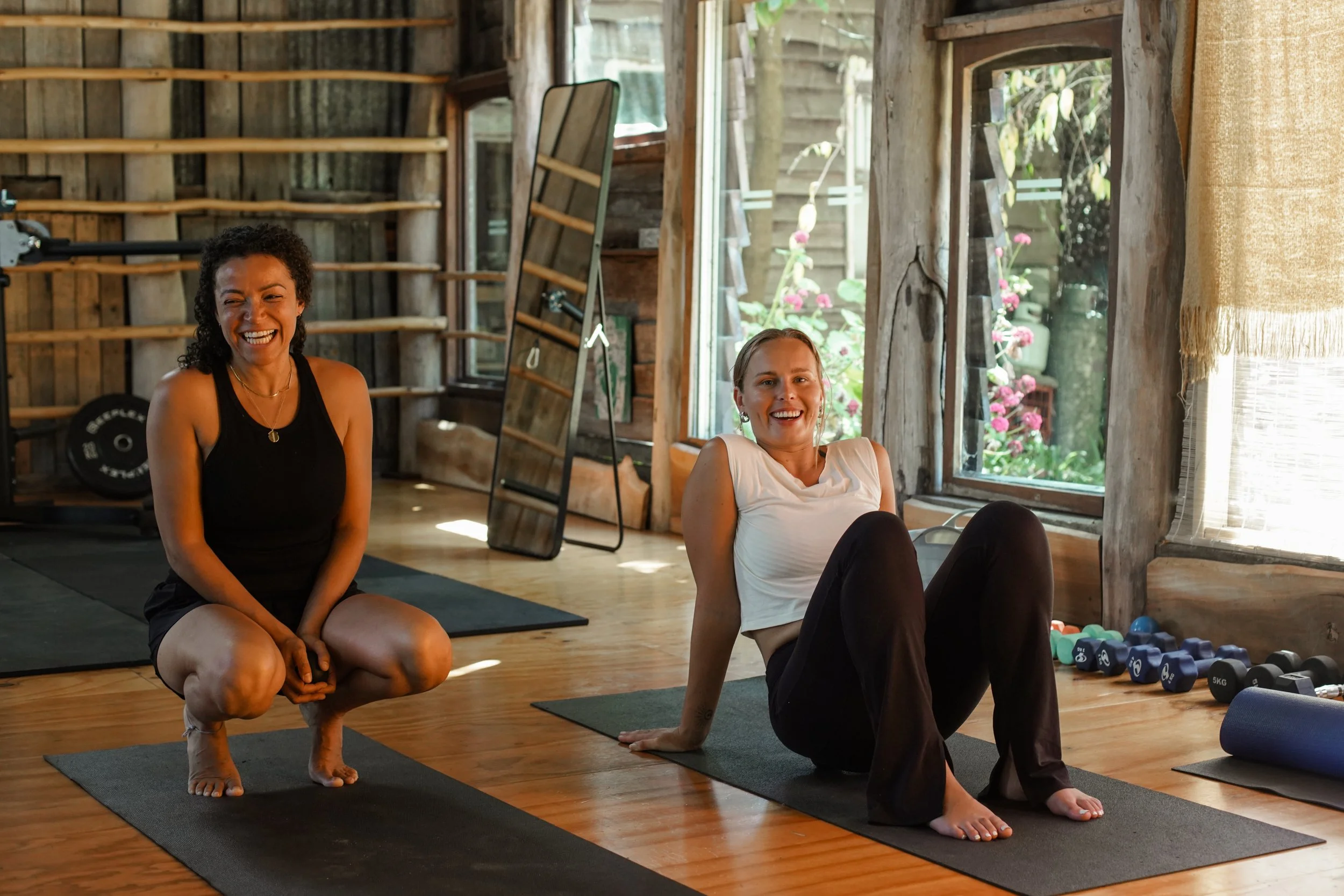 Two women exercising in a rustic wooden fitness studio with large windows, one sitting on a yoga mat and the other squatting, both smiling and enjoying their workout.