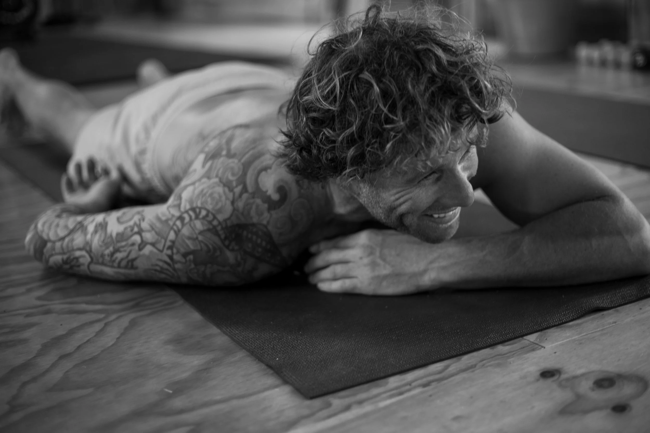 A man with curly hair and tattoos practicing yoga, lying face down on a yoga mat, smiling.