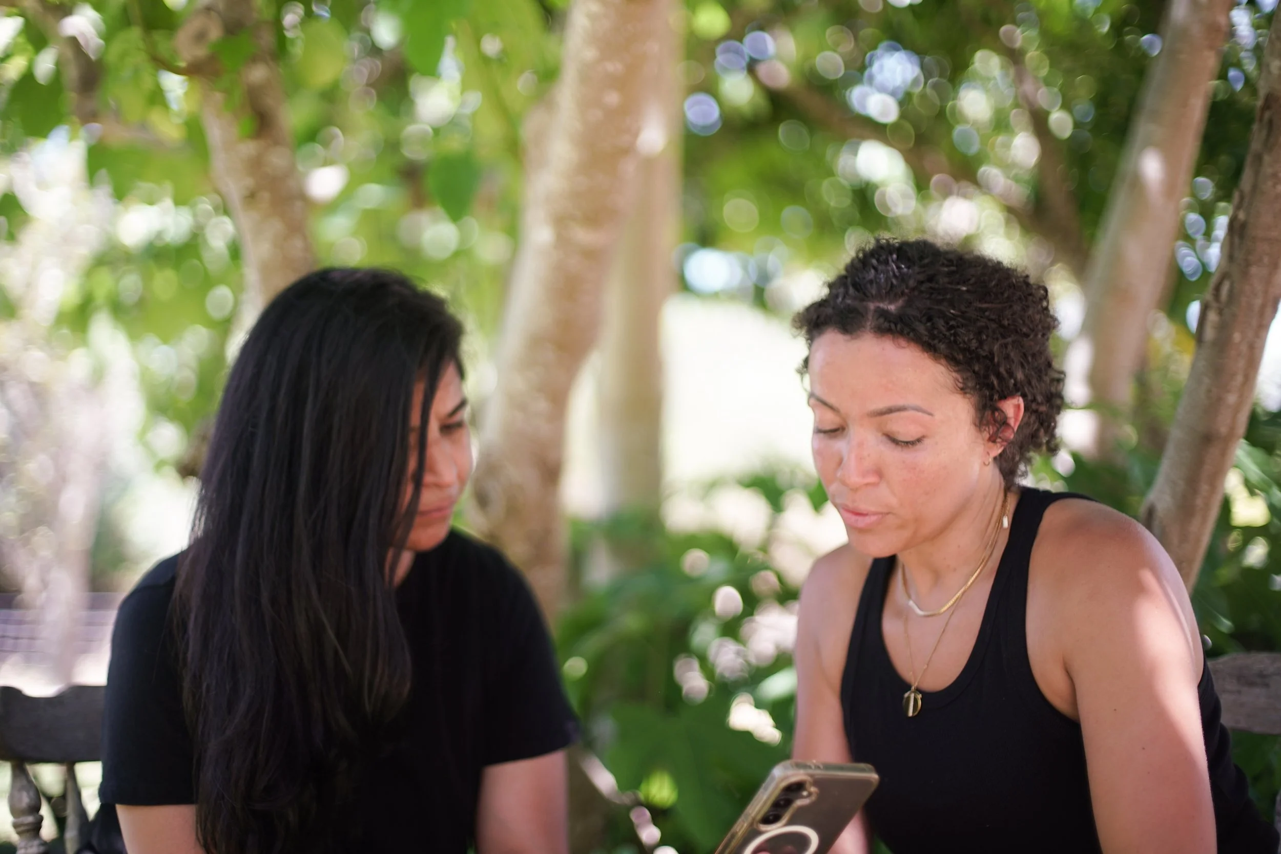 Two women sitting outdoors among trees, looking at a phone together.