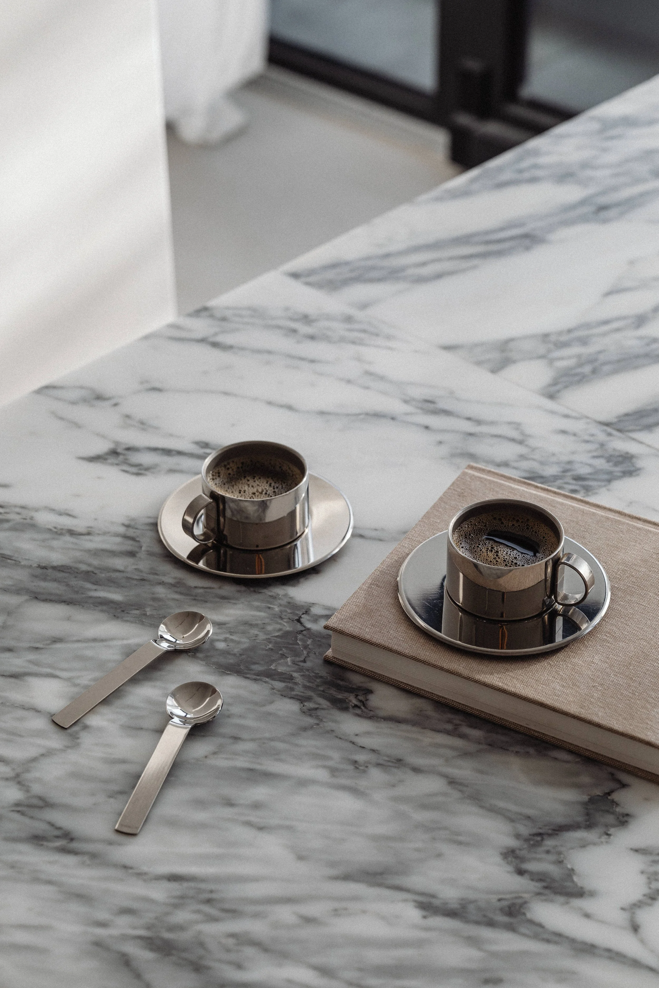 Two cups of coffee on metallic saucers placed on a white and gray marble table, with two silver teaspoons lying horizontally and a beige book