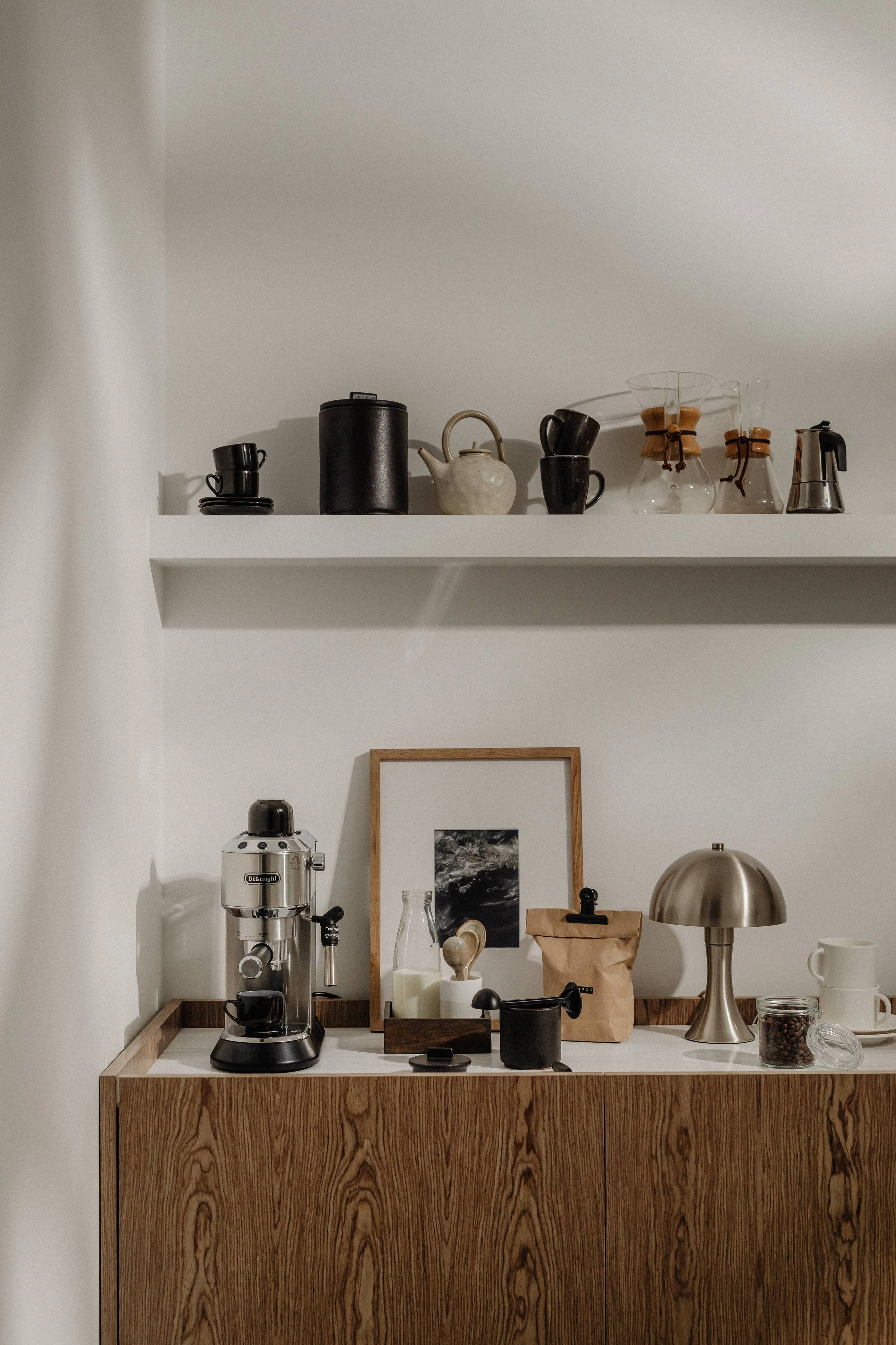 Minimalist kitchen counter with a silver espresso machine, a framed black and white photograph, a small milk bottle, a black stovetop, a paper bag, a metallic lamp, white cups, and coffee beans in a jar.
