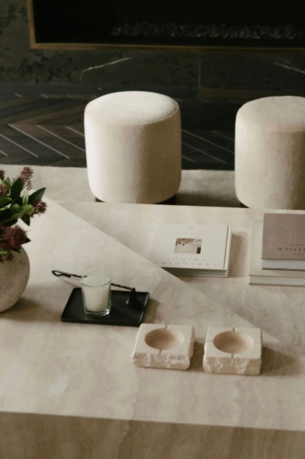 A table with books, a flower arrangement, a candle, and two white stone candle holders, with two white upholstered stools in the background.