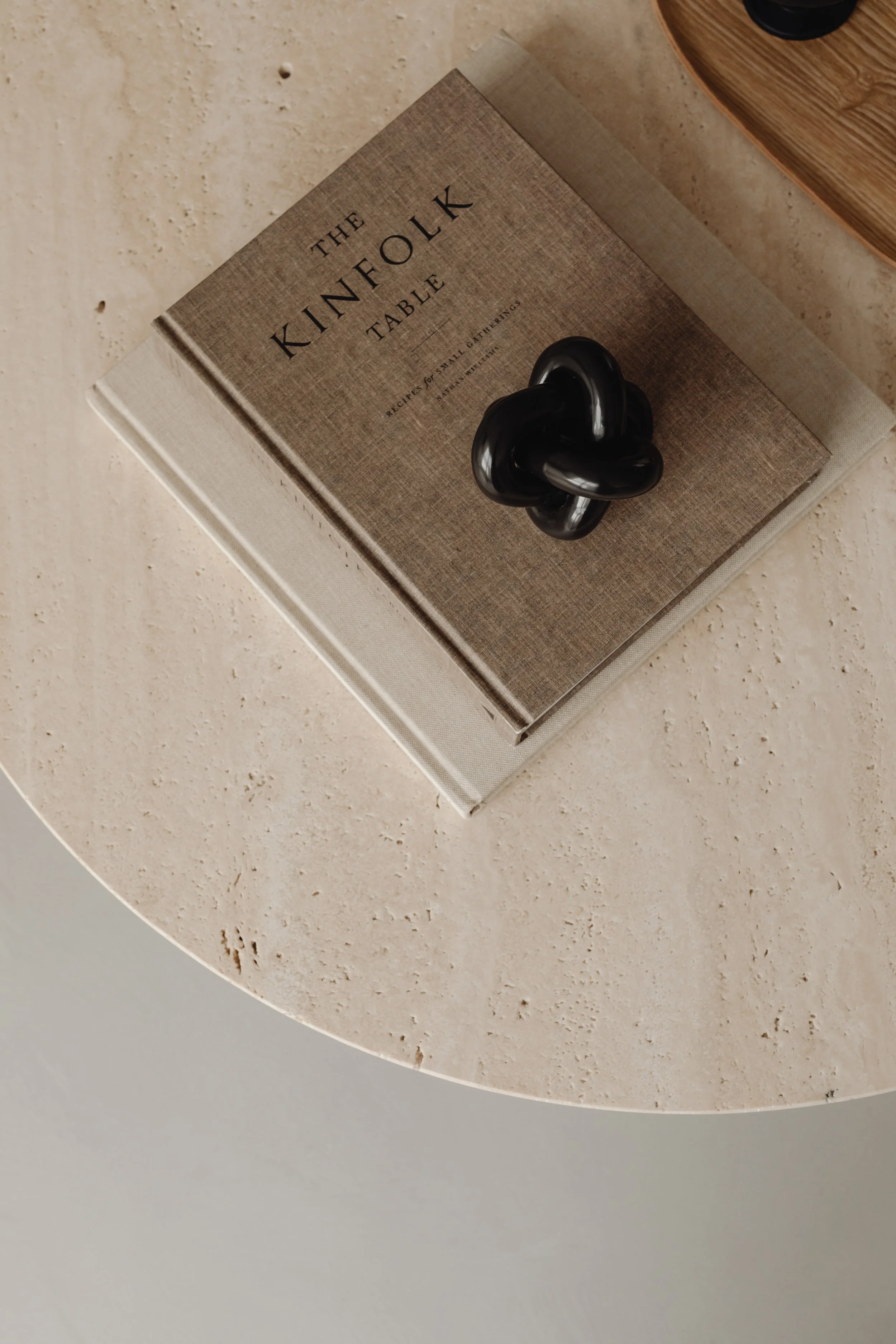 A hardcover book titled 'The Kinfolk Table' rests on a round, beige, textured table. A black, glossy sculpture is placed on top of the book.