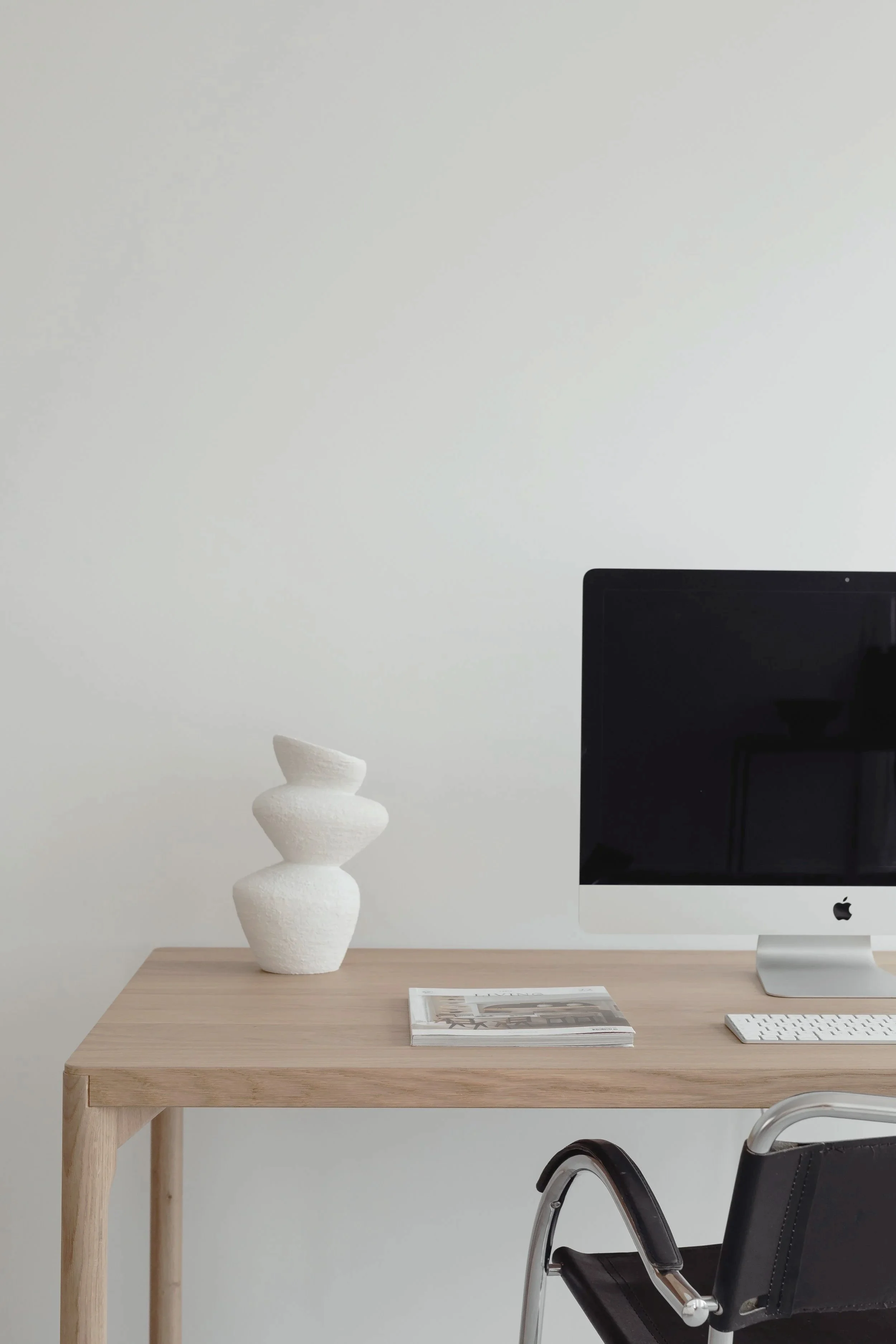 Minimalist workspace with a light wood desk, black office chair, white decorative vase with three stacked forms, a magazine, an iMac computer, and a white keyboard against a plain white wall.