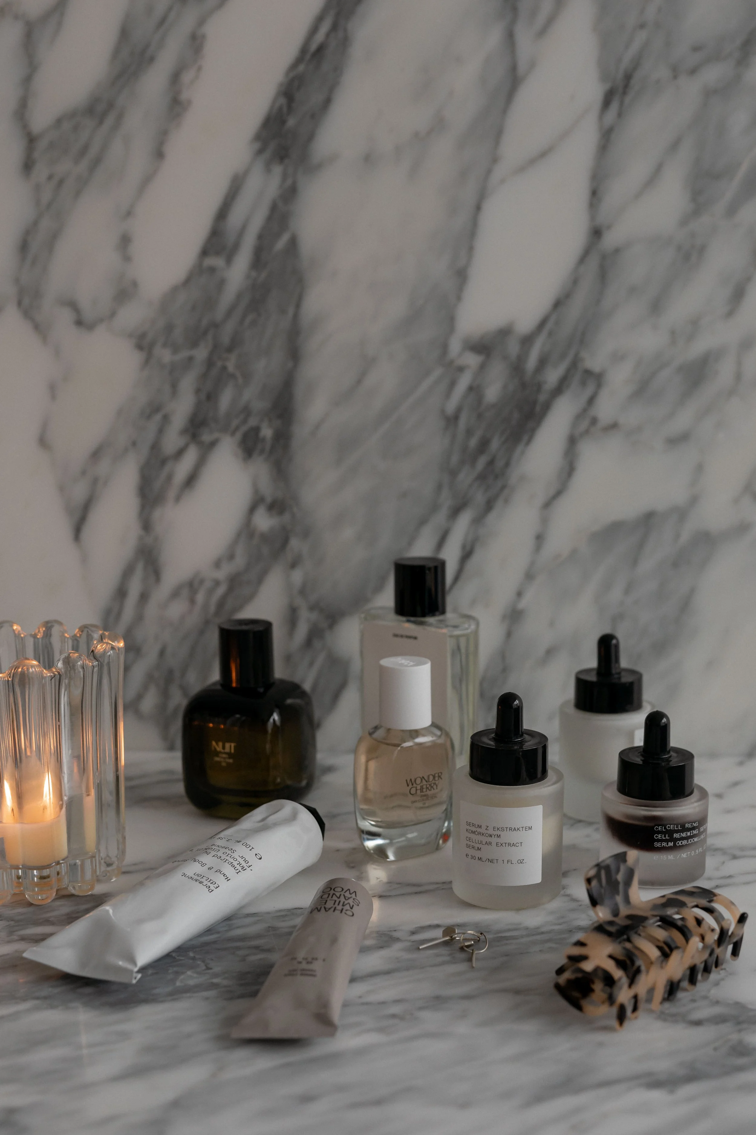 A collection of skincare products and accessories arranged on a marble surface, including bottles of serum, cream, and essential oils, with a lit candle in a glass holder and a tortoise shell hair clip.