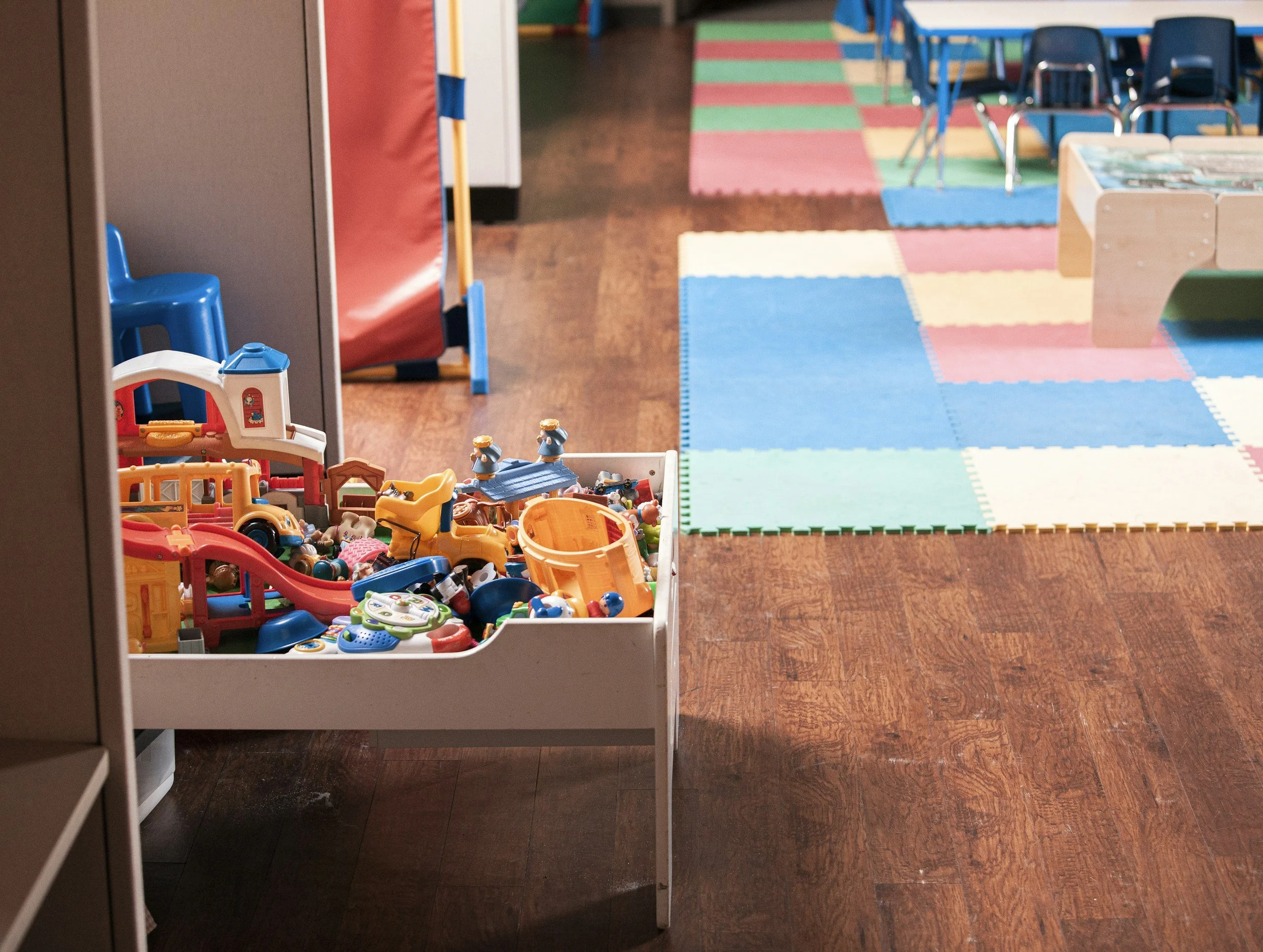 A children's play area with a white toy box filled with small plastic toys, including bulldozers, cars, and figurines, on a wooden floor. In the background, there are colorful foam mats and small chairs.