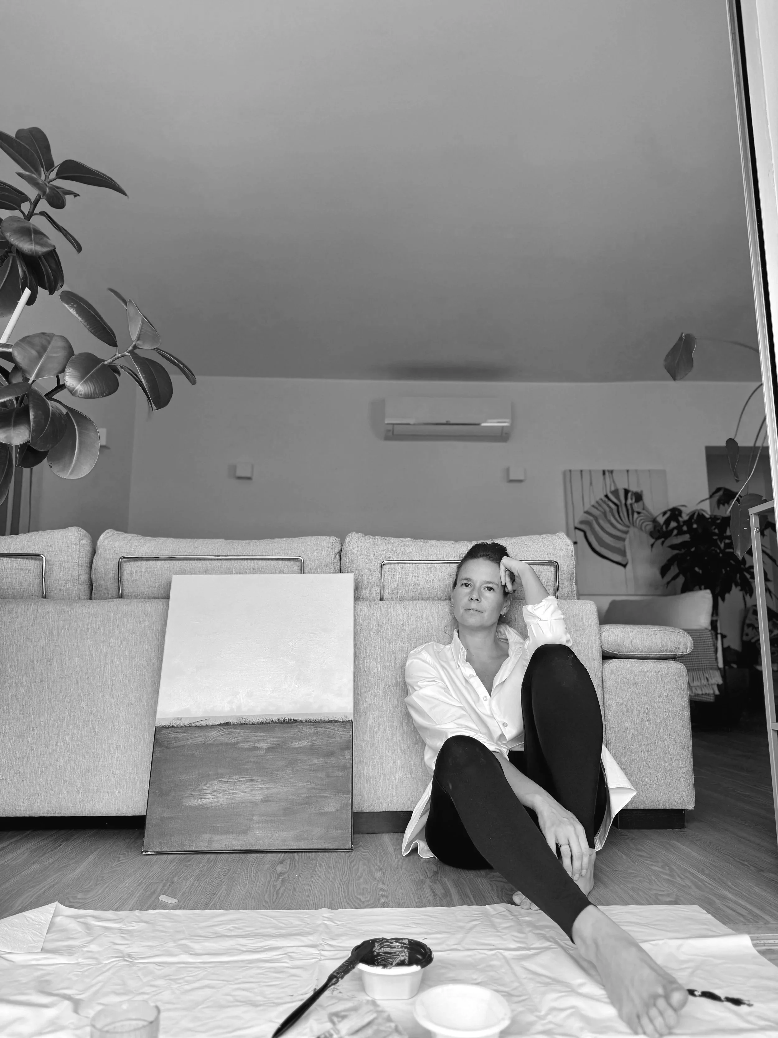 Mulher sentada no chão de frente para uma parede com um quadro de pintura ao lado, roupas e tintas ao redor, no ambiente de uma sala de estar.